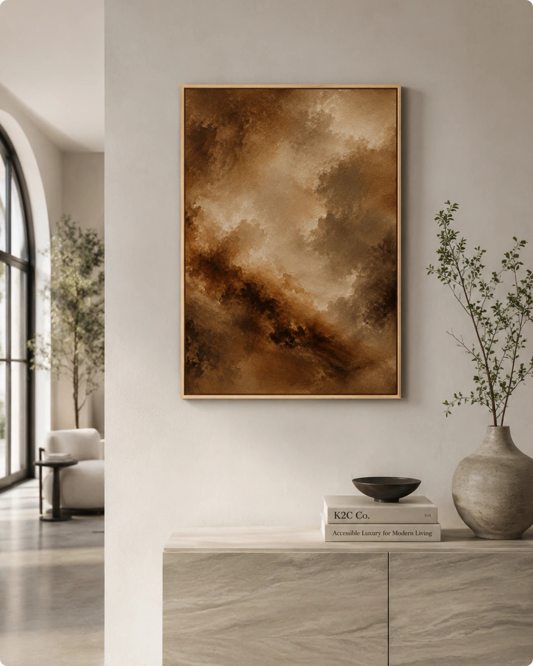 A framed abstract painting with warm brown, beige, and gold tones hangs on a light wall above a console table in a modern, sunlit living space.