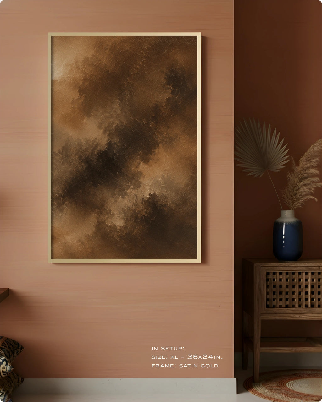 A framed abstract wall art print hangs on a warm terracotta wall in a styled interior. The artwork features a smoky, cloud-like texture in shades of brown, tan, and dark brown, enclosed in a thin satin gold frame.