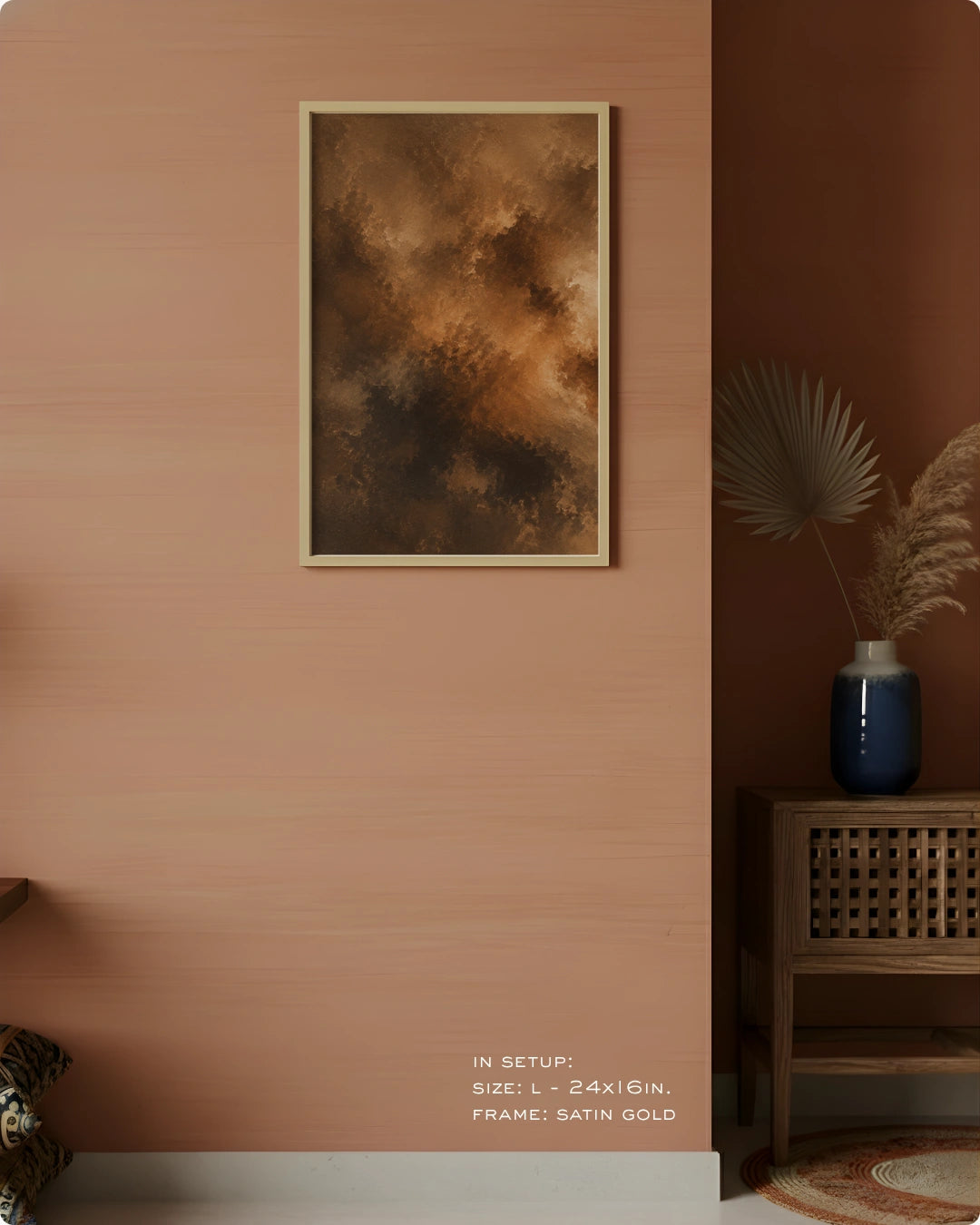 A framed abstract art print with warm brown, gold, and black tones hangs on a peach-colored wall in a styled interior setting with a wooden side table, blue vase, and dried palm leaves.