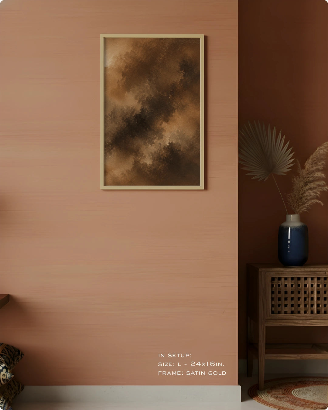 A framed abstract wall art print hangs on a warm peach-pink wall in a styled interior. The artwork features dark brown and tan cloud-like textures with a thin satin gold frame.
