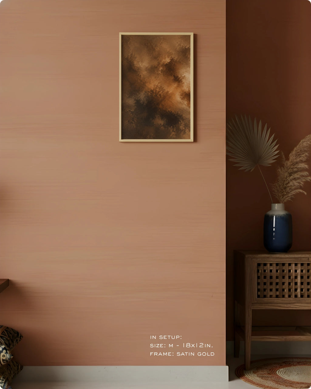 A framed abstract art print with a textured brown and gold design, hanging on a terracotta-colored wall in a styled interior setting with a wooden side table, blue vase, and dried palm leaves.