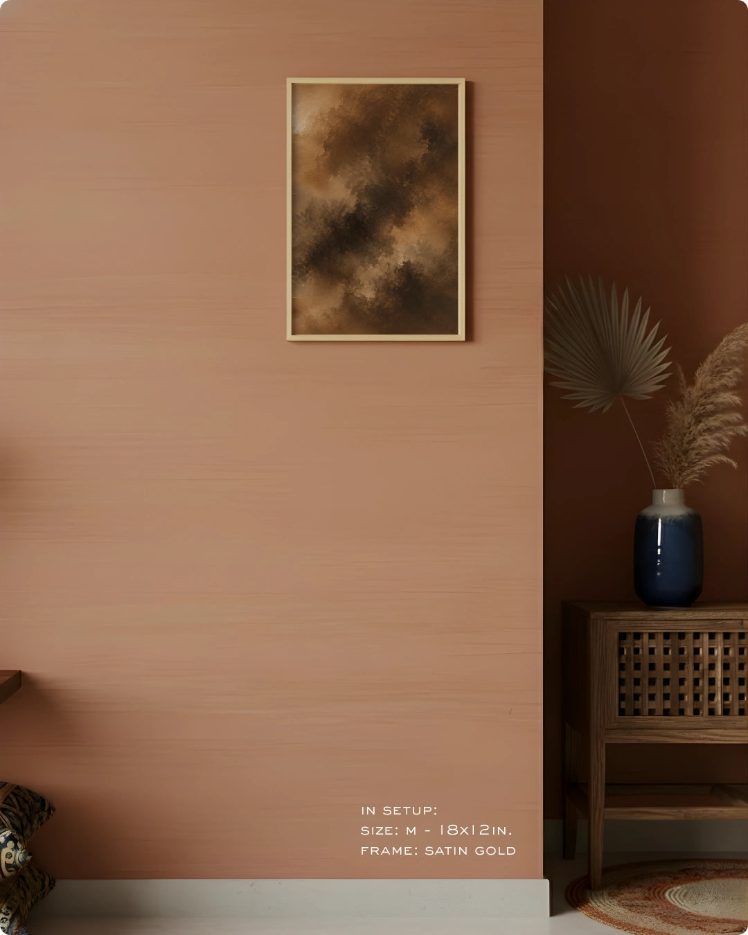 A framed abstract art print with a textured brown and gold design, hung on a warm terracotta-colored wall in a styled interior setting with a wooden side table and a blue vase.