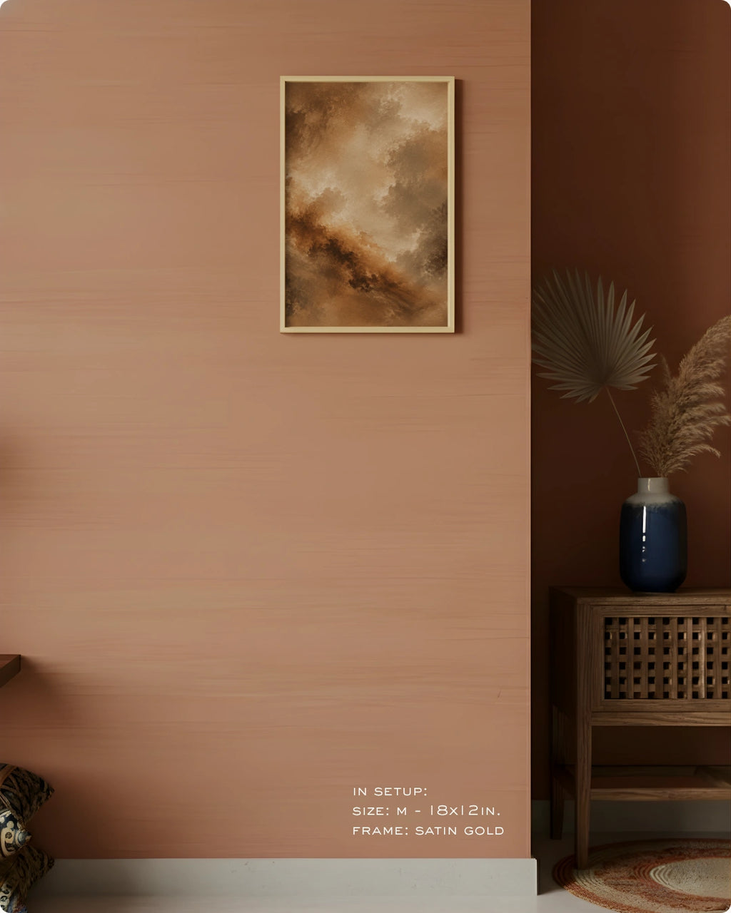 A framed abstract art print with warm brown, beige, and gold tones hangs on a terracotta-colored wall in a styled interior setting with a wooden side table, blue vase, and dried palm leaves.