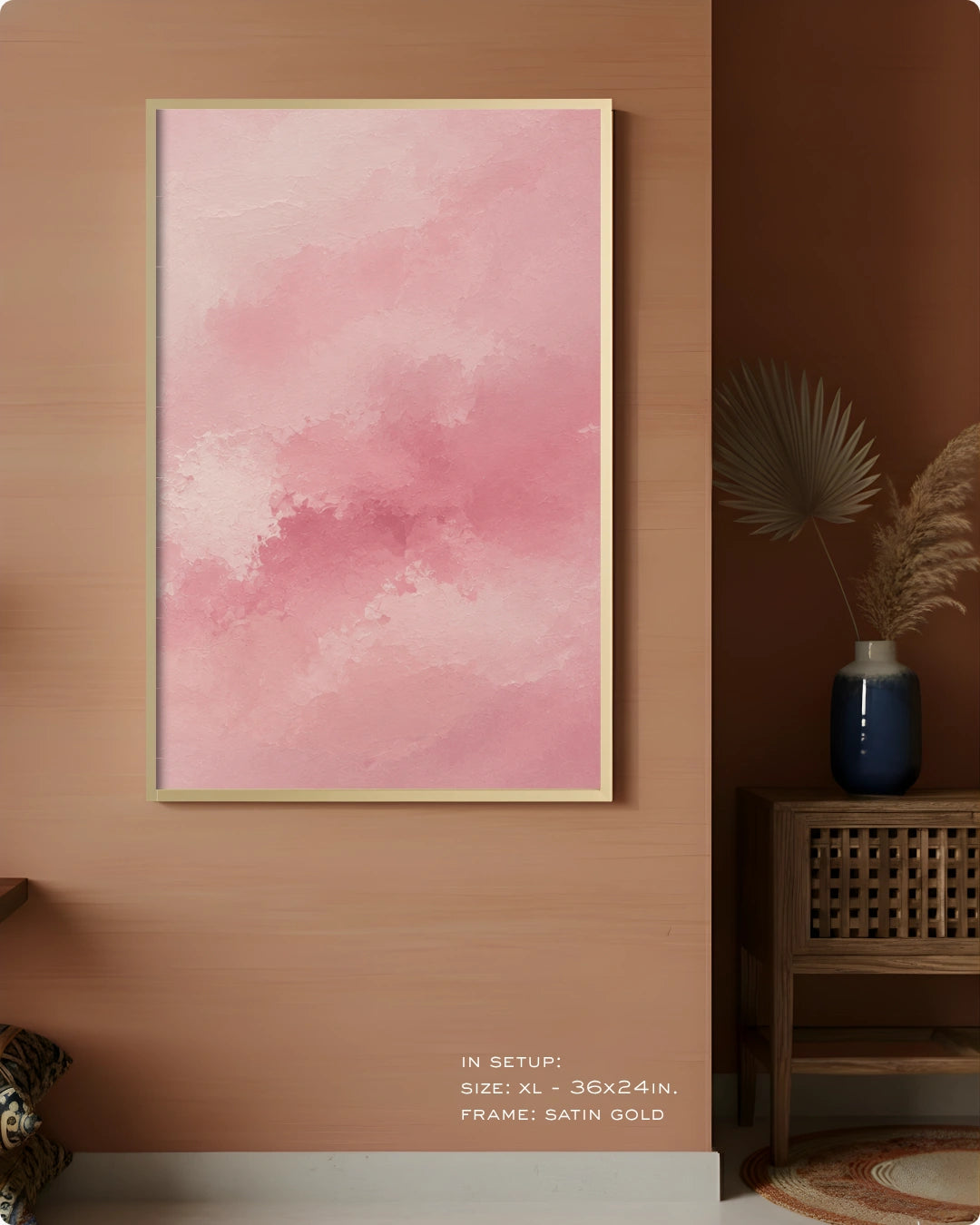 A vertical framed wall art print with a soft pink watercolor texture, displayed on a warm wood-paneled wall in a styled interior setting with a side table and vase.