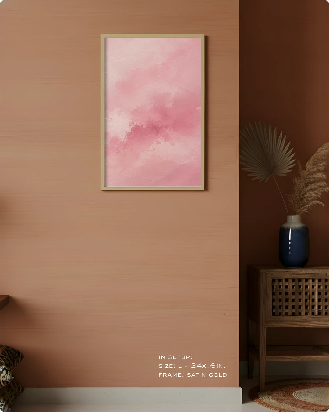 A framed abstract pink watercolor art print hanging on a warm peach wall in a styled interior setting with a wooden side table, blue vase, dried palm leaves, and a small rug. Text at the bottom reads: IN SETUP: STEEL S = 24K GOLD, FRAME: SATIN GOLD.