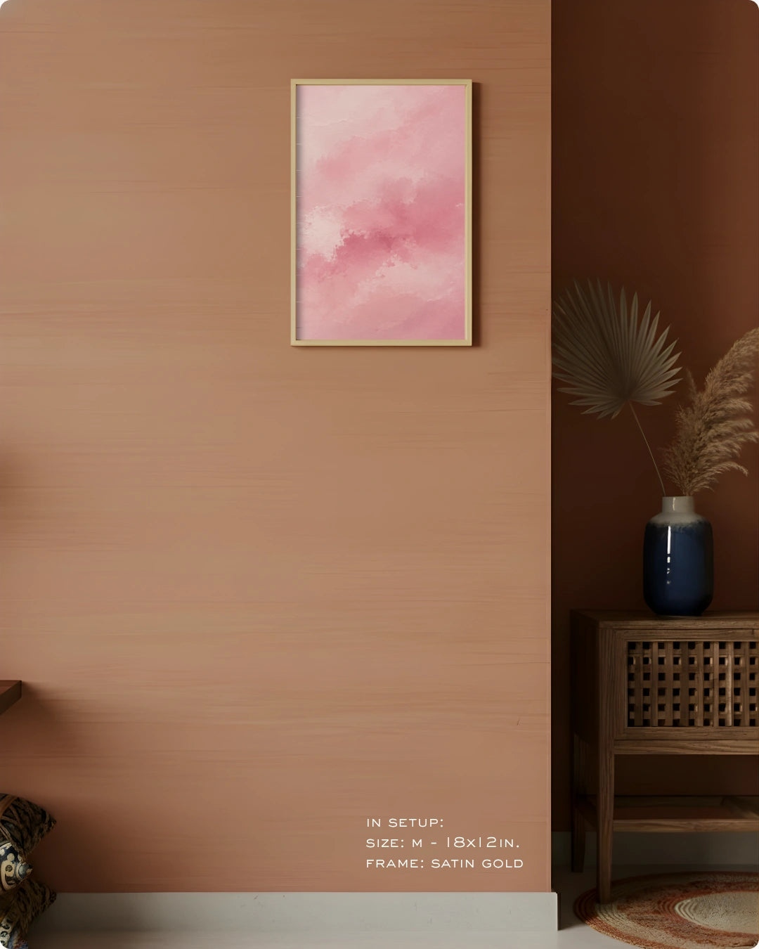 A framed wall art print with a soft pink watercolor design hangs on a warm terracotta-colored wall in a styled interior setting. The frame is satin gold, and the background includes a wooden side table, a blue vase with dried palm leaves, and a dark brown corner wall.