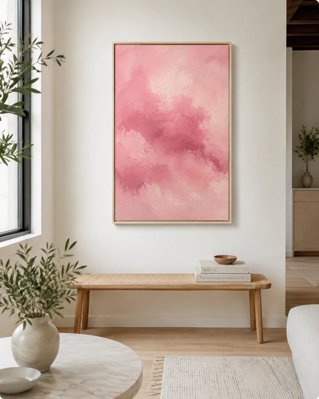 A framed abstract pink painting hangs on a white wall above a light wood bench in a bright, modern living room with plants and a marble coffee table.