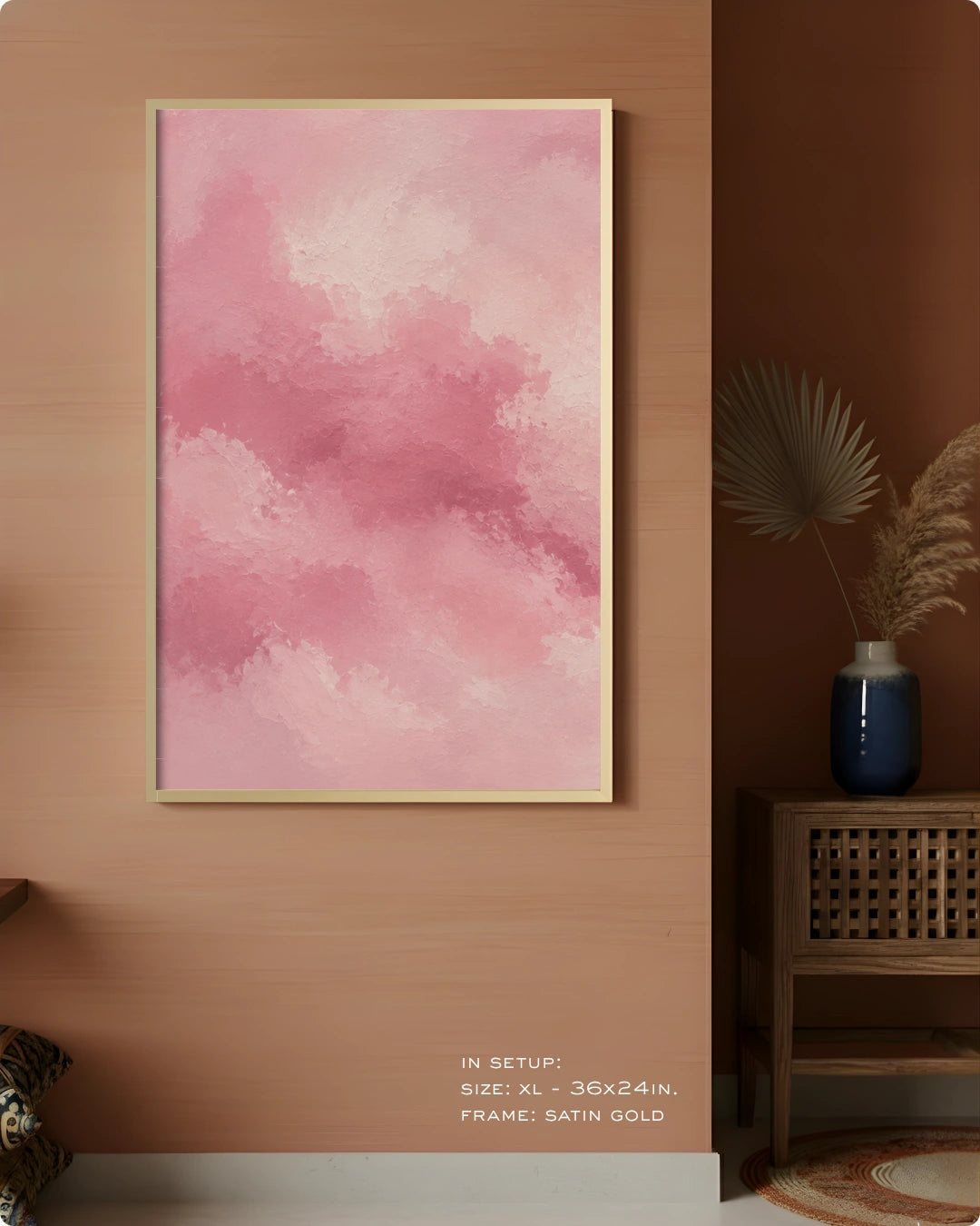 A vertical framed wall art print with a soft pink watercolor cloud design, displayed on a warm wood wall in a styled interior setting with a side table and vase.