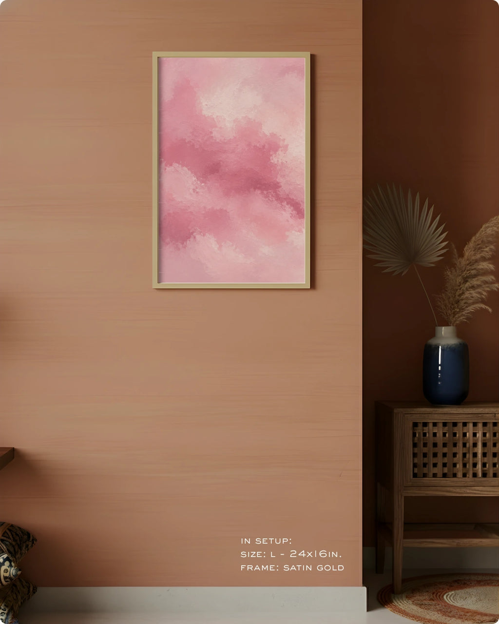 A framed wall art print with a pink watercolor cloud design hangs on a warm peach-colored wall in a styled interior setting. The frame is satin gold, and the background includes a wooden side table, a blue vase with dried grass, and a dark brown corner wall.