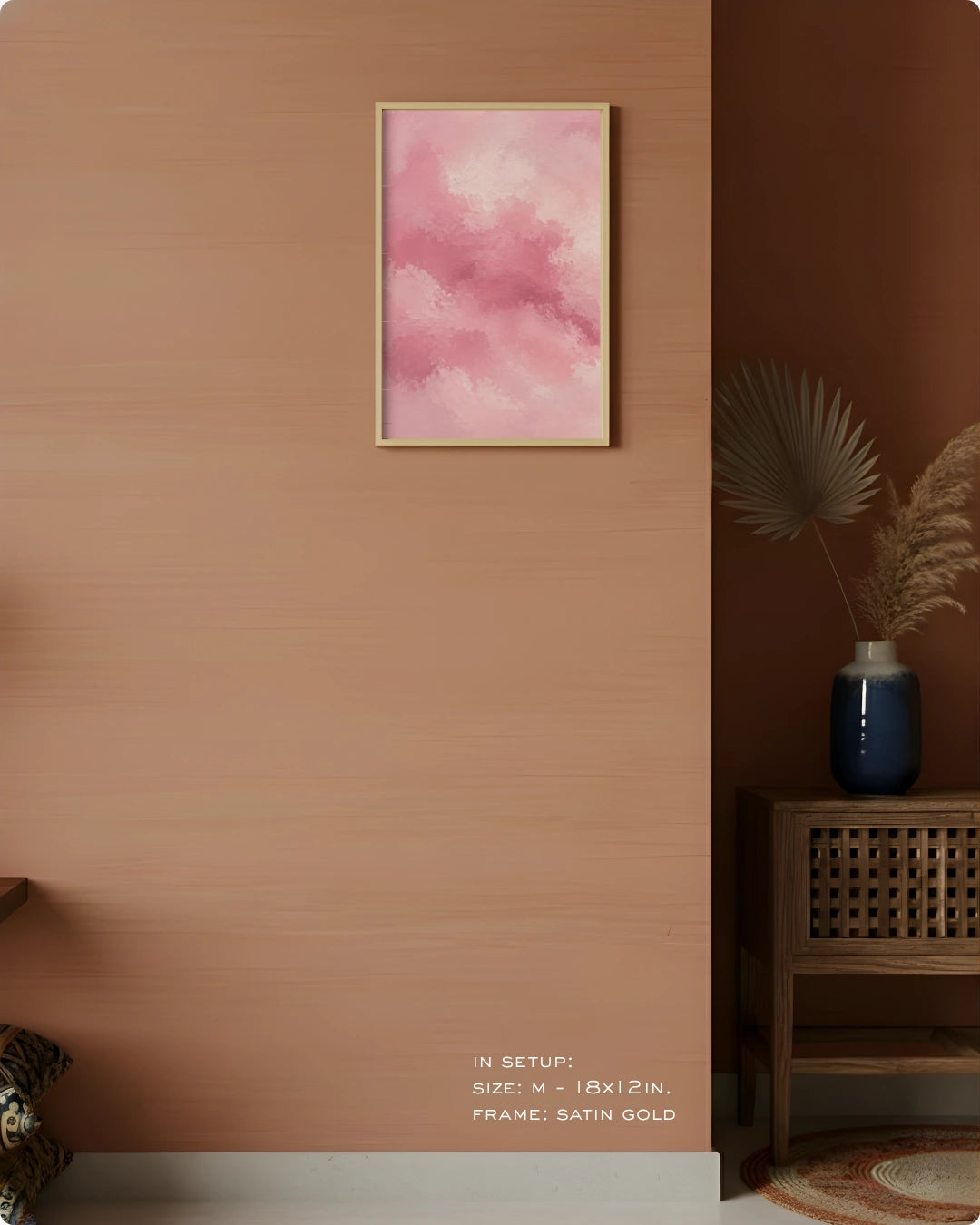 A framed abstract pink watercolor art print hangs on a warm terracotta wall in a styled interior setting with a wooden side table, blue vase, dried palm leaves, and a small rug. Text at the bottom reads 'IN SETUP: SIZE - 18x24, FRAME - SATIN GOLD.