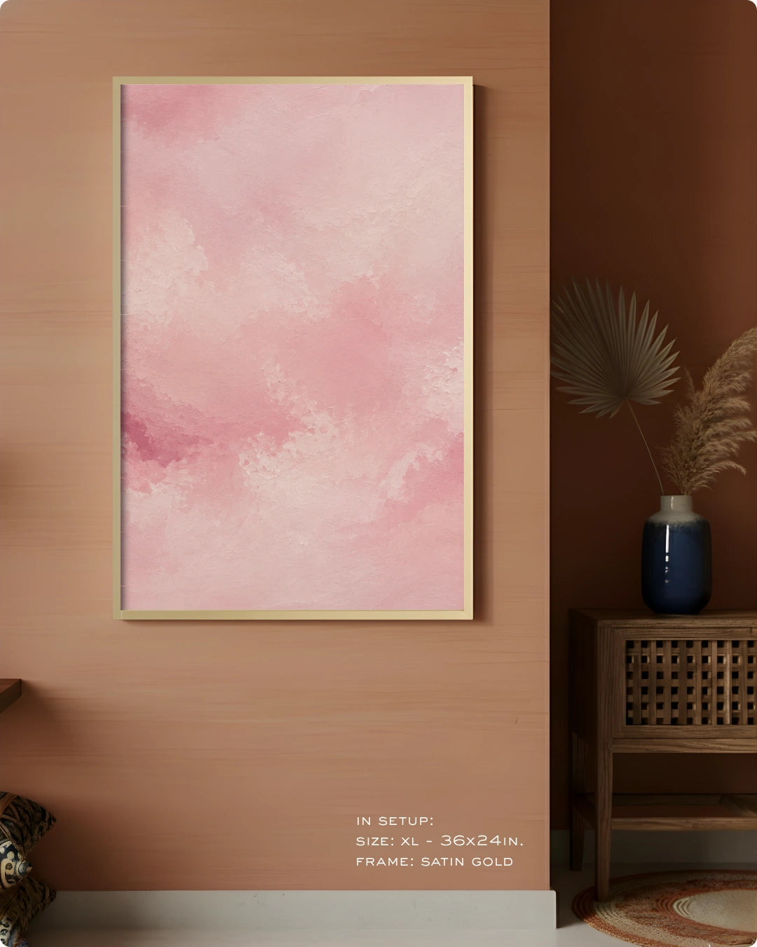 A vertical framed wall art print with a soft pink watercolor texture hangs on a warm wood wall in a styled interior. The frame appears satin gold, and the room includes a side table, blue vase, dried palm leaves, and a small decorative object.