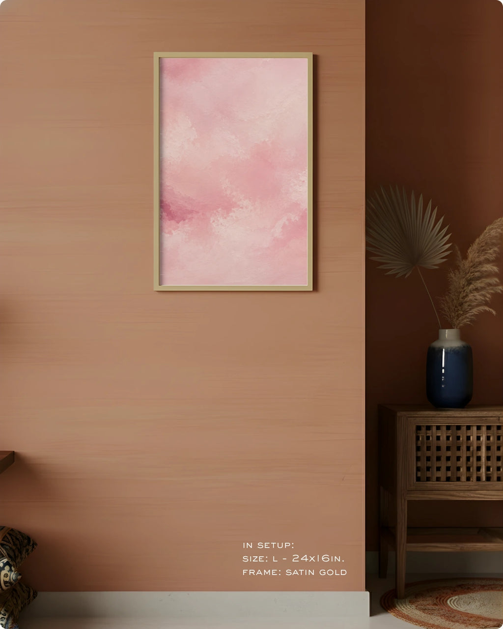 A framed abstract art print with soft pink watercolor clouds hangs on a warm peach-colored wall in a styled interior setting. The frame is satin gold.