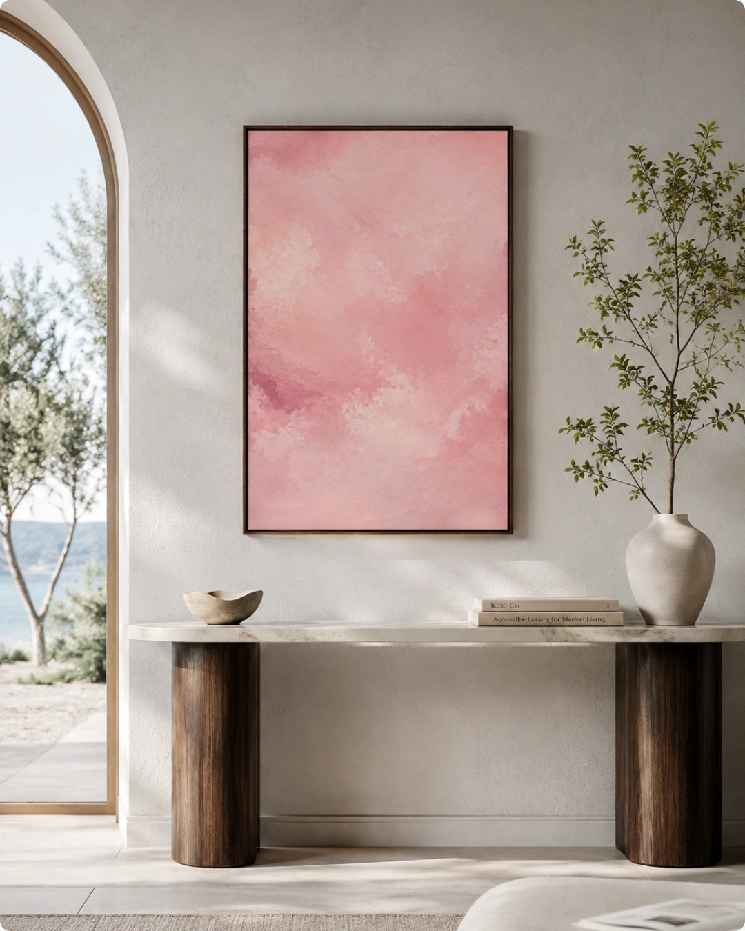 A framed abstract pink painting hangs on a light beige wall above a console table in a styled interior setting with a vase, books, and an arched window view.