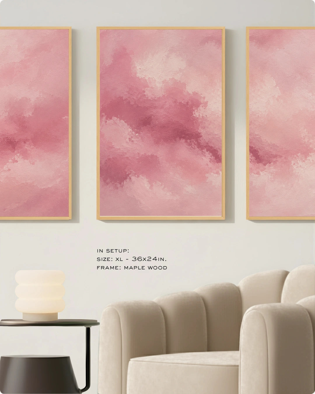 Three vertical framed wall art panels with soft pink watercolor-style abstract designs, displayed above a cream sofa in a styled interior. Small text reads: 'IN SETUP: SIZE: XL - 36x24in, FRAME: MAPLE WOOD'.