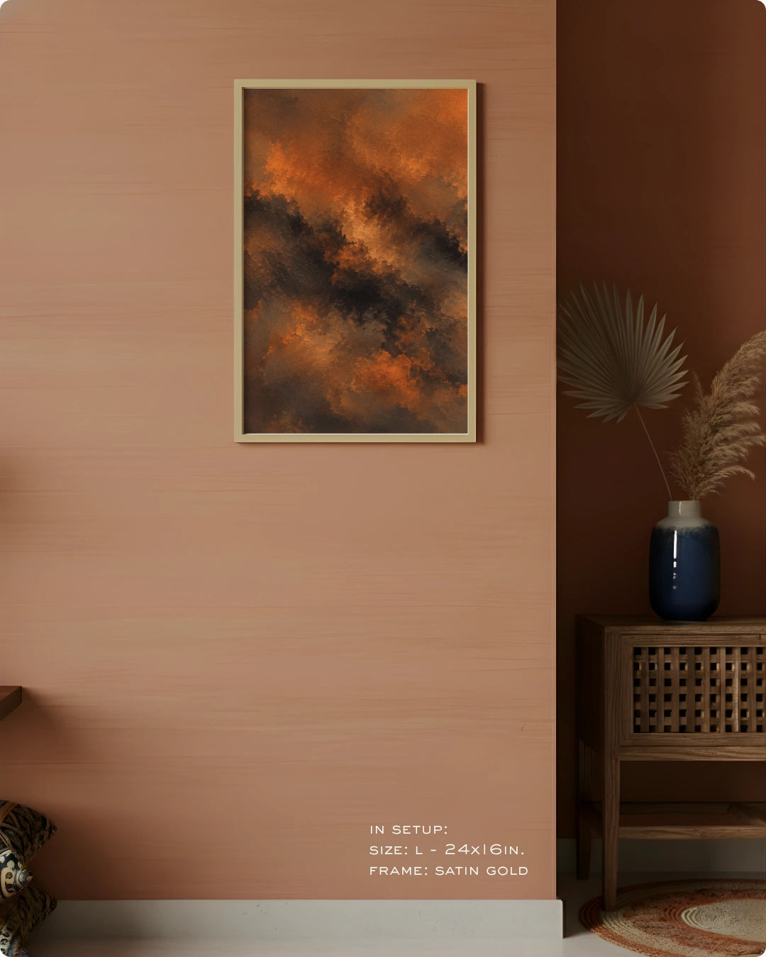 A framed abstract wall art print hangs on a peach-colored wall in a styled interior. The artwork features swirling orange, brown, and dark charcoal tones, set in a satin gold frame.