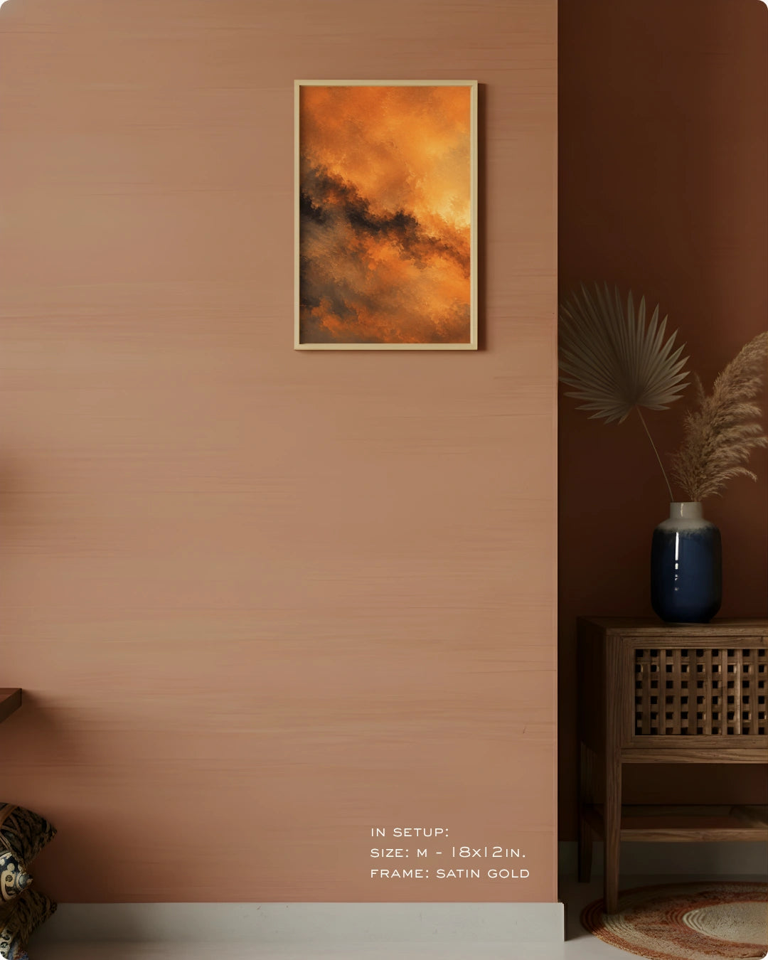 A framed abstract art print featuring vibrant orange and brown clouds hangs on a warm terracotta-colored wall in a styled interior setting with a wooden side table, blue vase, and dried palm leaves.