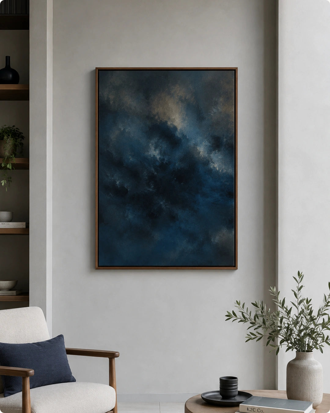 Lifestyle mockup of the moody abstract art in a walnut frame styled above a side table next to a minimalist cream lounge chair with a navy pillow.