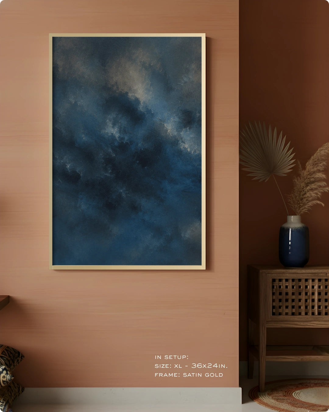 A vertical framed wall art print showing a moody dark blue cloudy sky, displayed on a warm terracotta wall in a styled interior setting with a side table and vase.