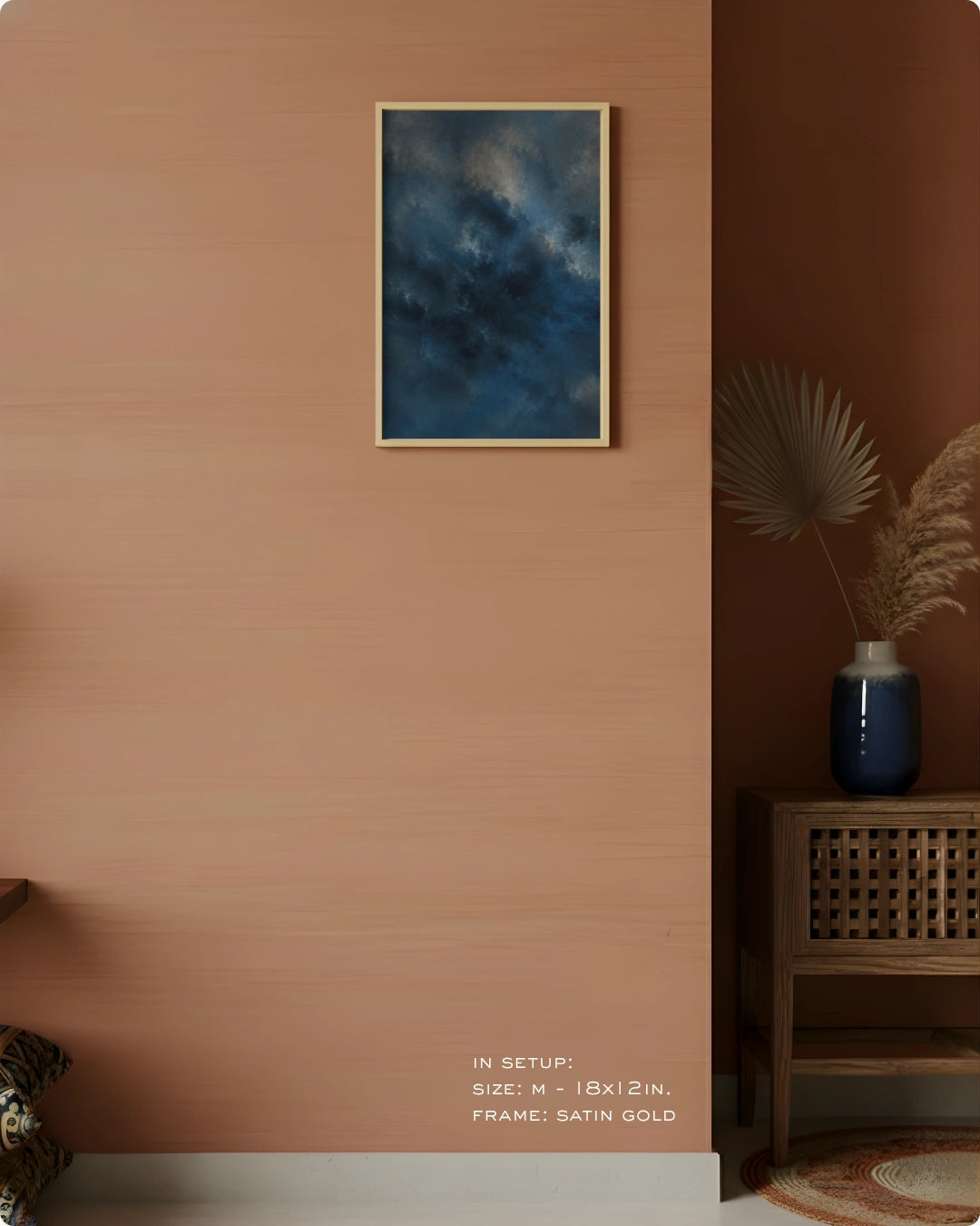 A framed wall art print depicting a dark blue cloudy sky, styled in a modern interior setting with a warm terracotta wall, a wooden side table, and a blue vase.