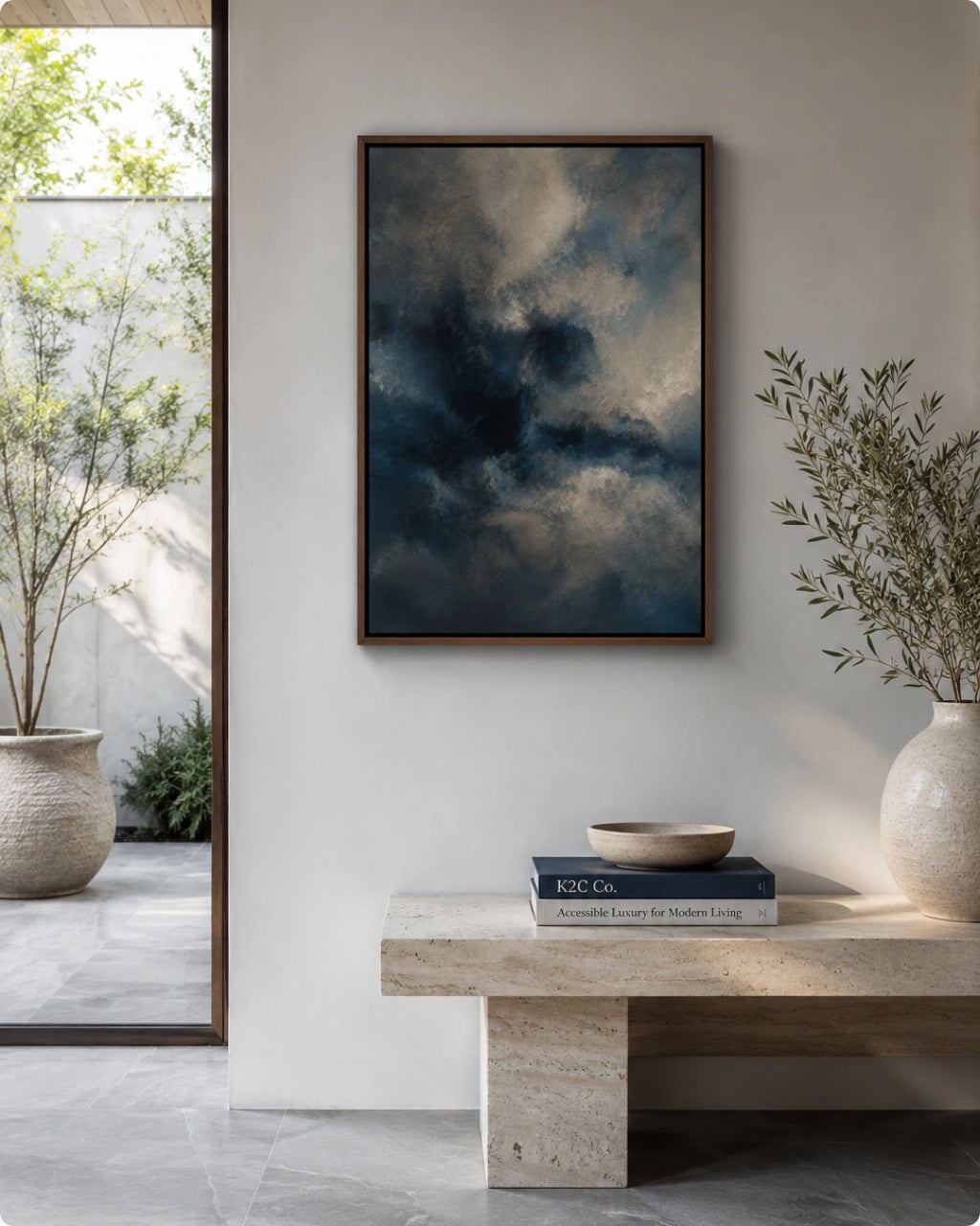 A framed vertical art print depicting a moody, dark blue and gray cloudy sky, displayed on a light gray wall above a stone bench in a modern, sunlit interior.