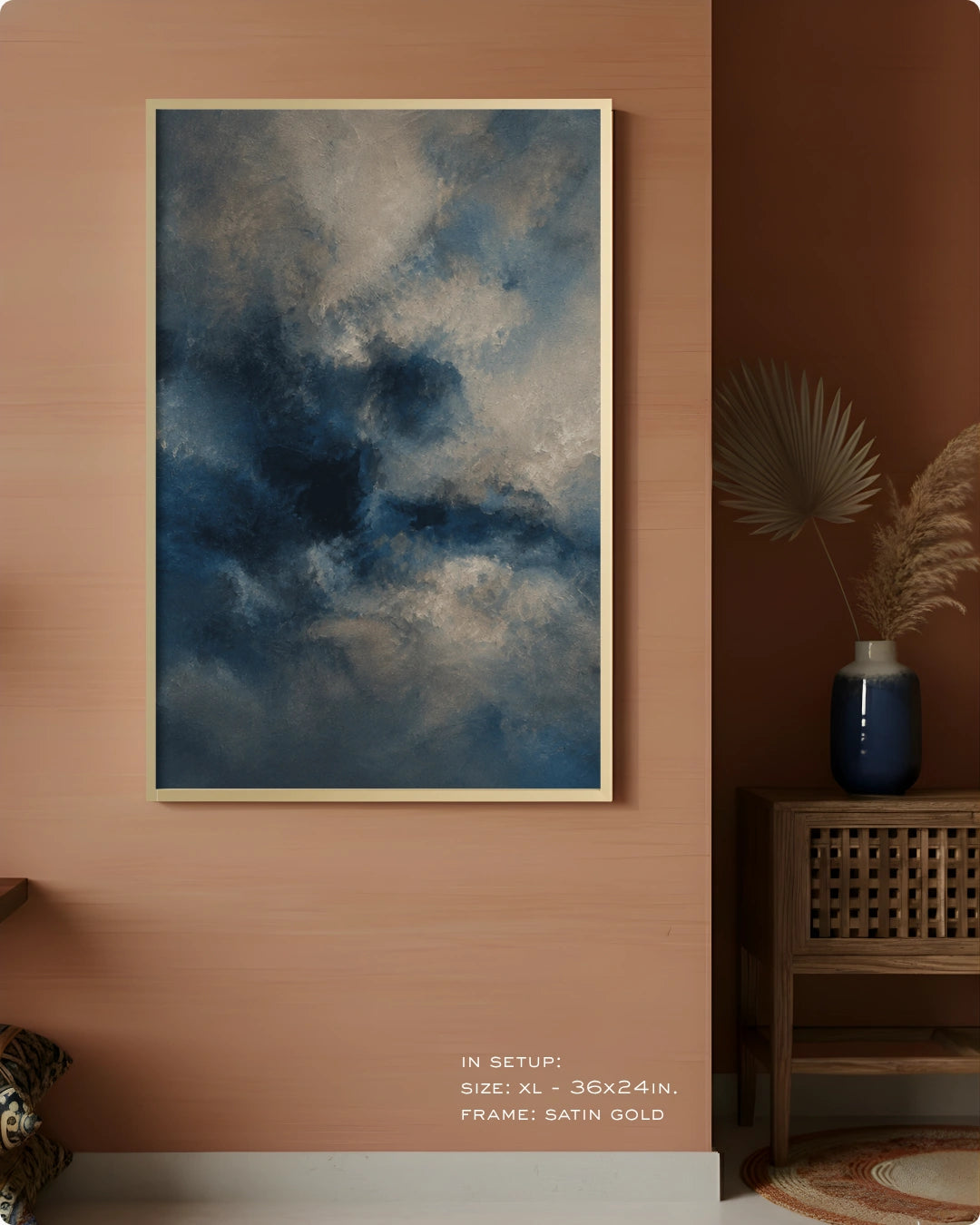 A framed wall art print showing a dramatic cloudy sky in deep blue, gray, and white tones, displayed on a warm terracotta wall in a styled interior setting with a side table and vase.