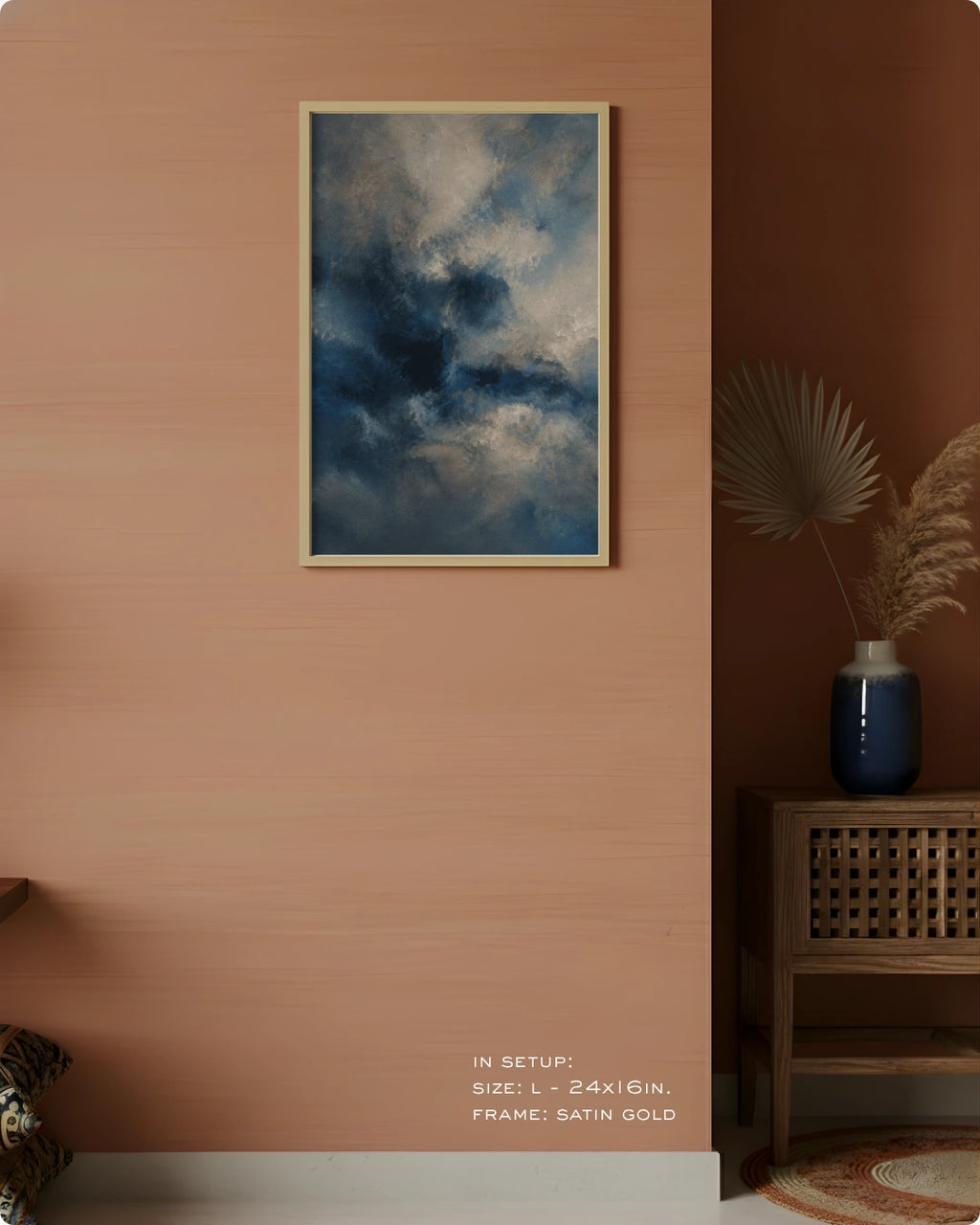 A framed wall art print depicting a dramatic blue and gray cloudy sky, hung on a warm peach-colored wall in a styled interior setting with a wooden side table, blue vase, and dried palm leaves.