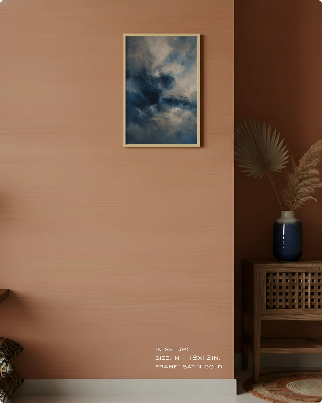 A framed wall art print depicting a dramatic blue and white stormy sky, hung on a warm terracotta-colored wall in a styled interior setting with a wooden side table, blue vase, and dried palm leaves.
