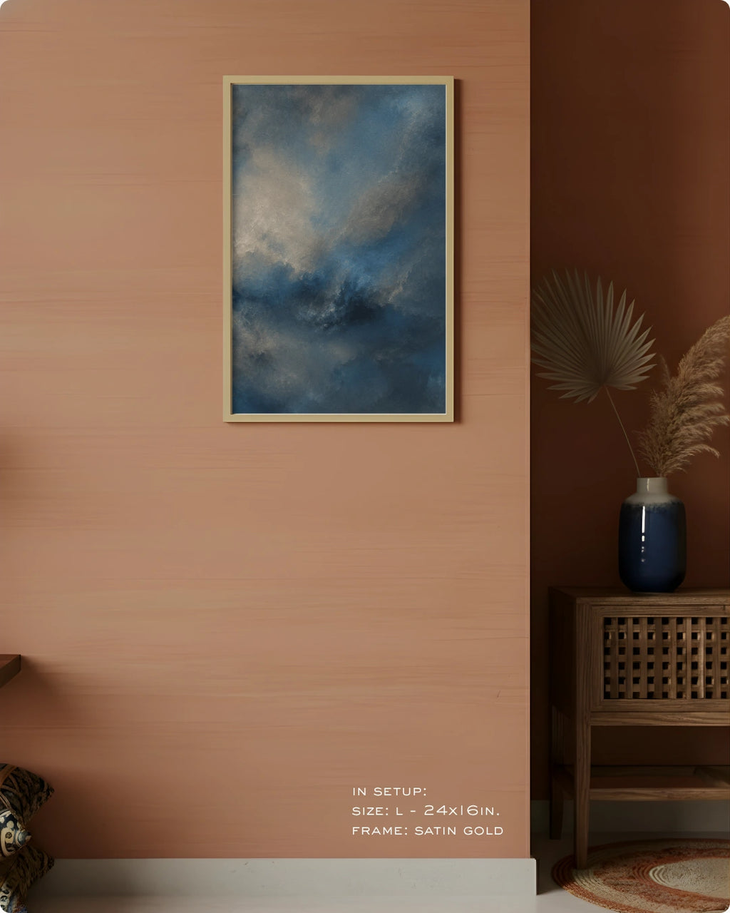 A framed wall art print featuring a dramatic blue and gray cloudy sky, displayed in a vertical orientation on a warm peach-colored wall. The frame is satin gold.