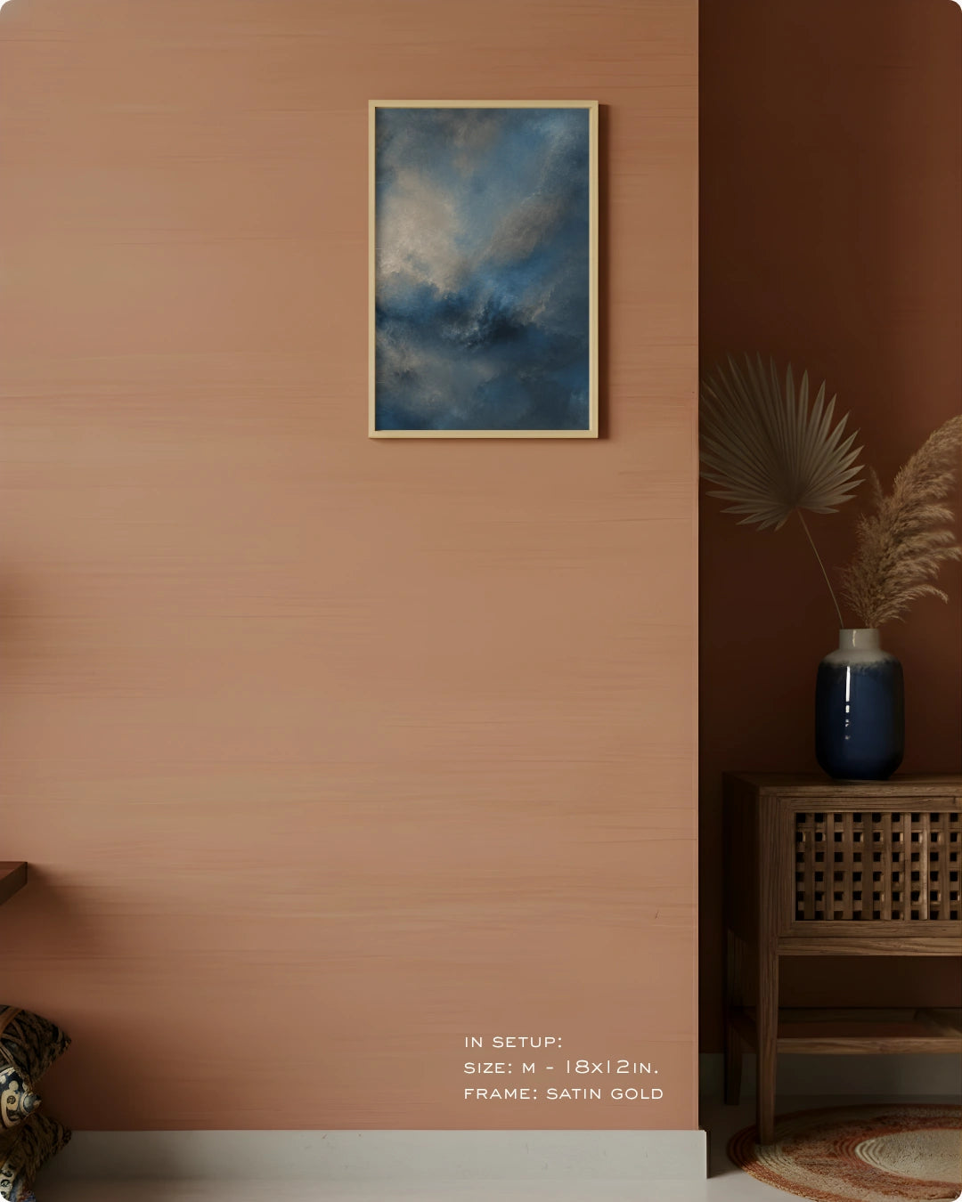 A framed wall art print depicting a dramatic blue and gray cloudy sky, hung on a warm terracotta-colored wall in a styled interior setting with a wooden side table, blue vase, and dried palm leaves.