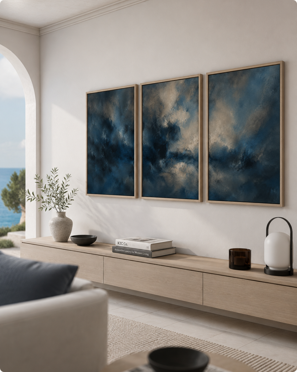 Three framed abstract canvas prints with deep blue and gray cloud-like imagery, displayed in a light wood frame on a white wall above a modern light wood console in a styled living room.