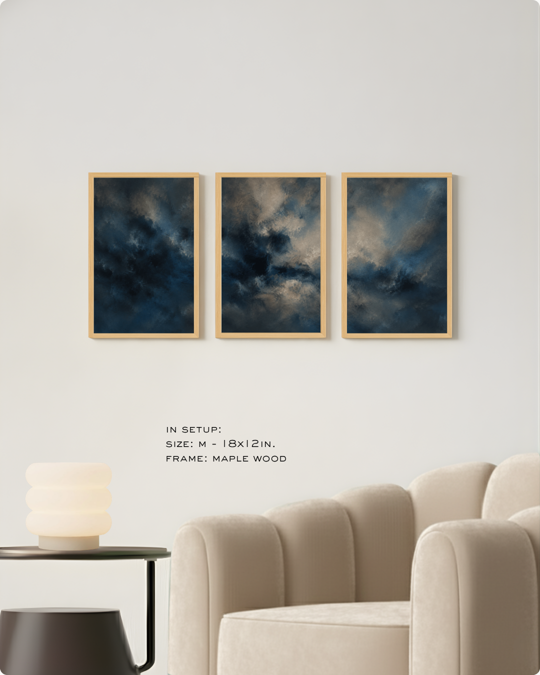 Three framed abstract wall art prints with dark blue and gray storm-like clouds hang on a light wall above a beige armchair. Small text reads 'IN SETUP: SIZE: M - 18x12 IN.