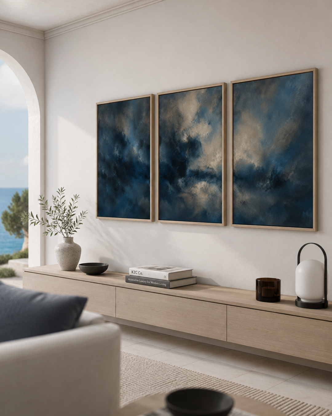 Three framed abstract canvas prints with deep blue and gray cloud-like imagery, displayed in a light wood frame on a white wall above a modern light wood console in a styled living room.