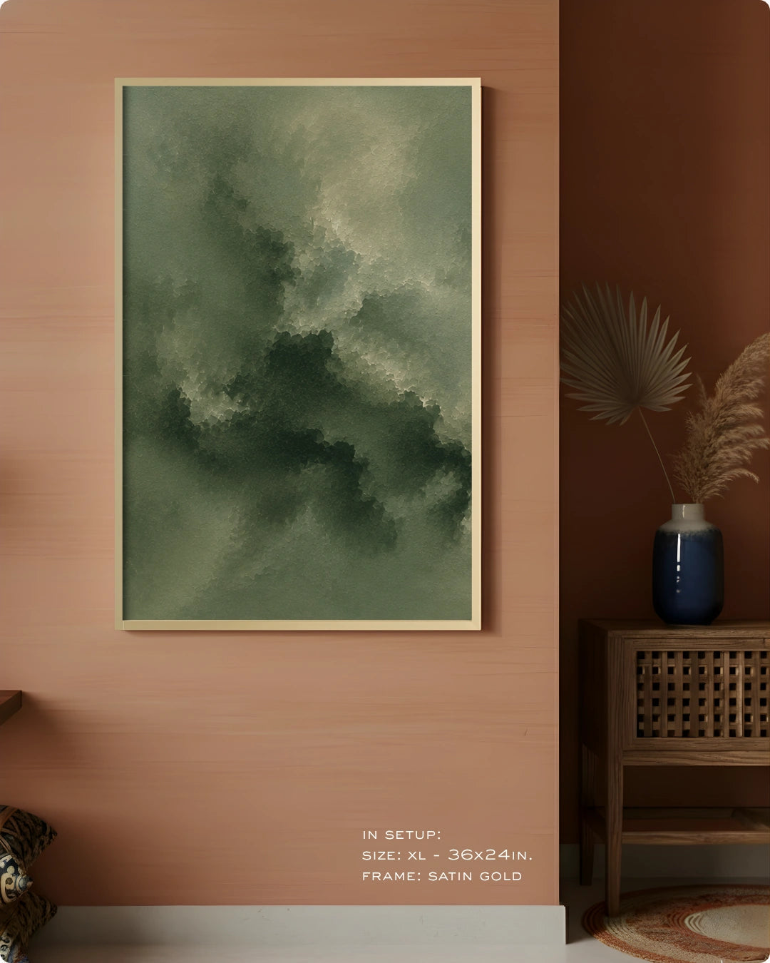 A framed abstract wall art print hangs on a warm terracotta wall in a styled interior. The artwork features a textured, painterly composition in muted green and gray tones with a thin gold frame.