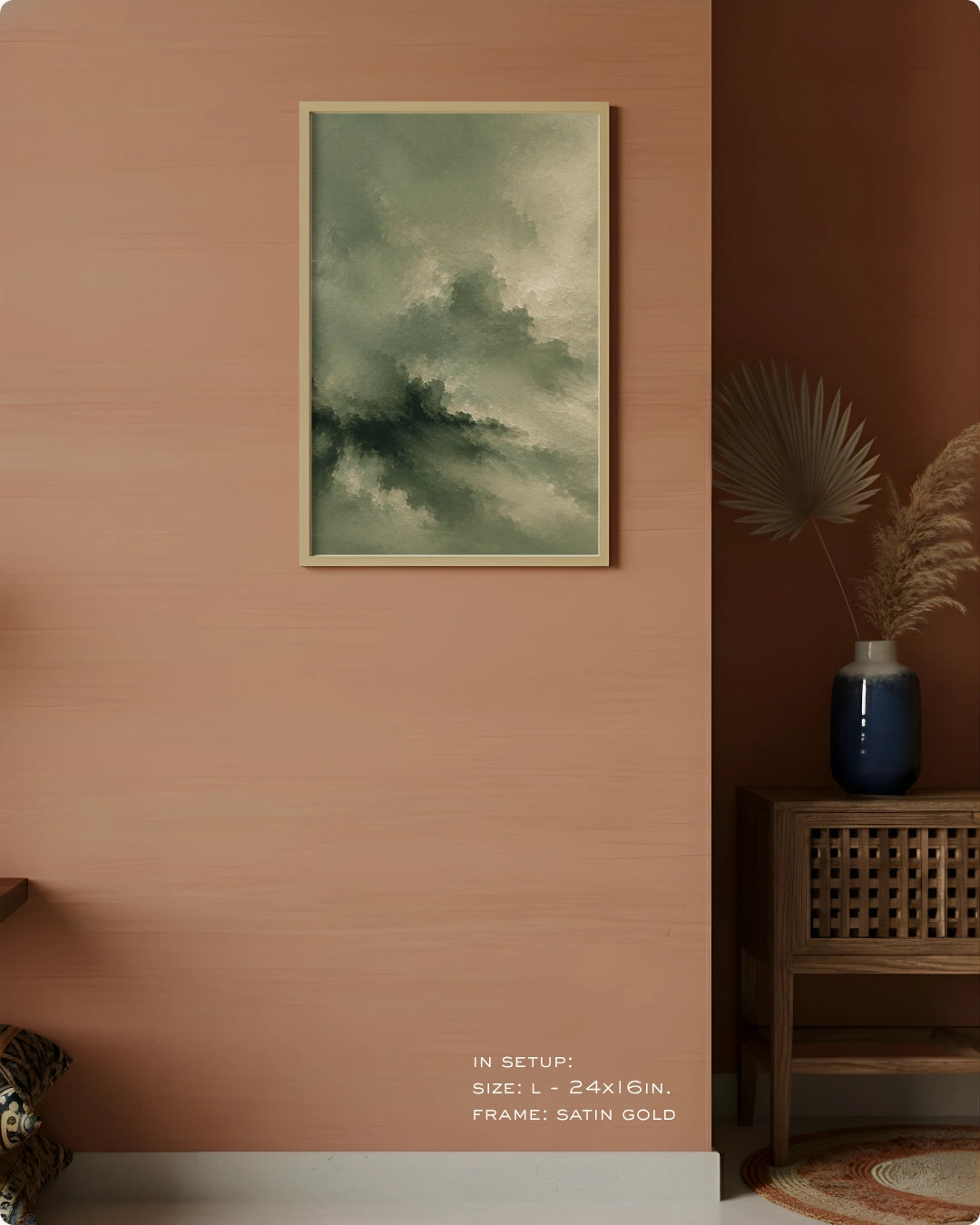A framed abstract landscape art print featuring green and gray clouds, displayed on a textured terracotta wall in a styled interior setting with a wooden side table and a blue vase.