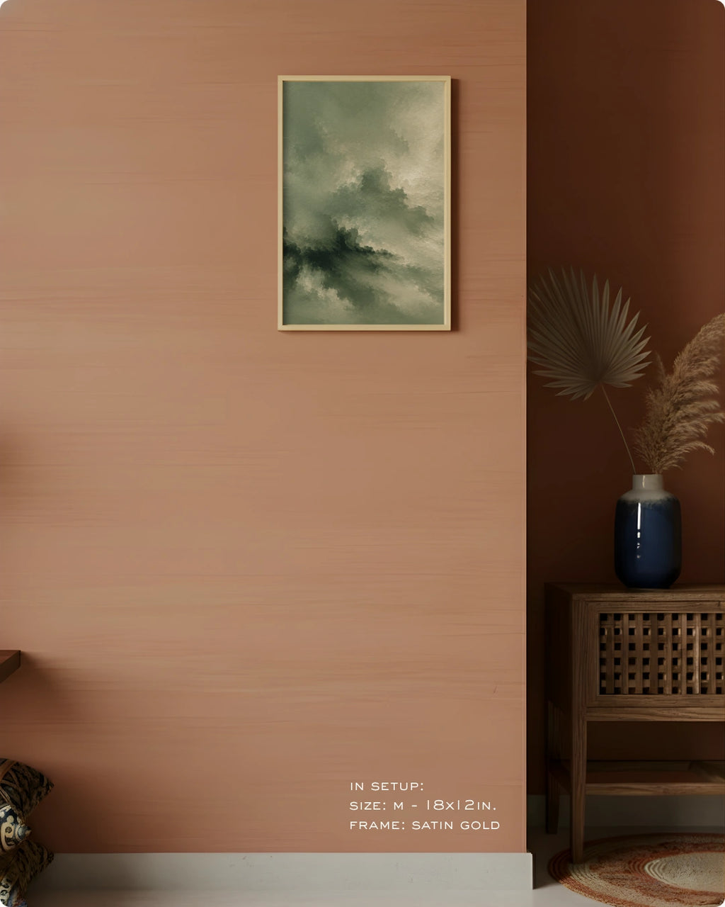 A framed abstract landscape art print featuring green and gray clouds, displayed on a textured terracotta wall in a styled interior setting with a wooden side table and a blue vase.