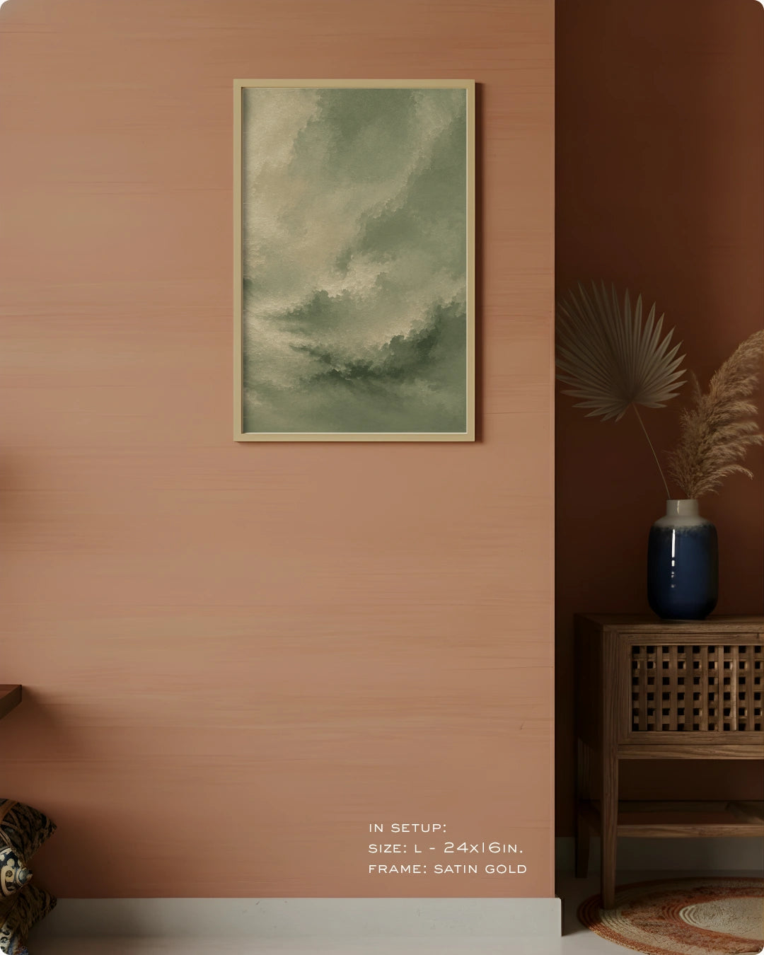 A framed vertical art print with a muted green and gray cloudy design, displayed on a textured terracotta wall in a styled interior setting with a wooden side table, blue vase, and dried palm leaves.