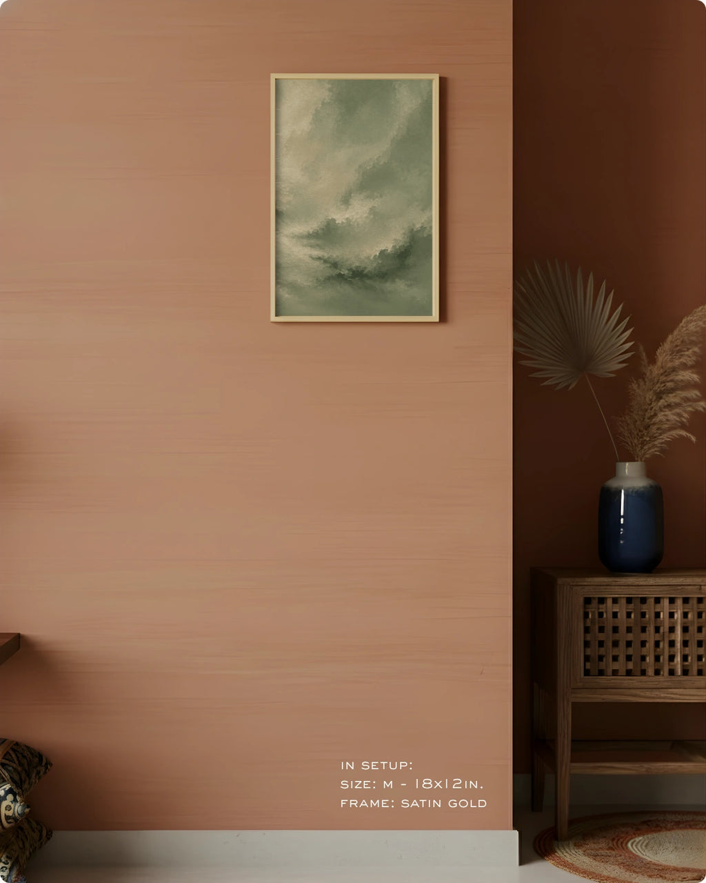 A framed art print depicting a stormy green sky hangs on a warm terracotta-colored wall in a styled interior setting with a wooden side table, blue vase, and dried palm leaves.
