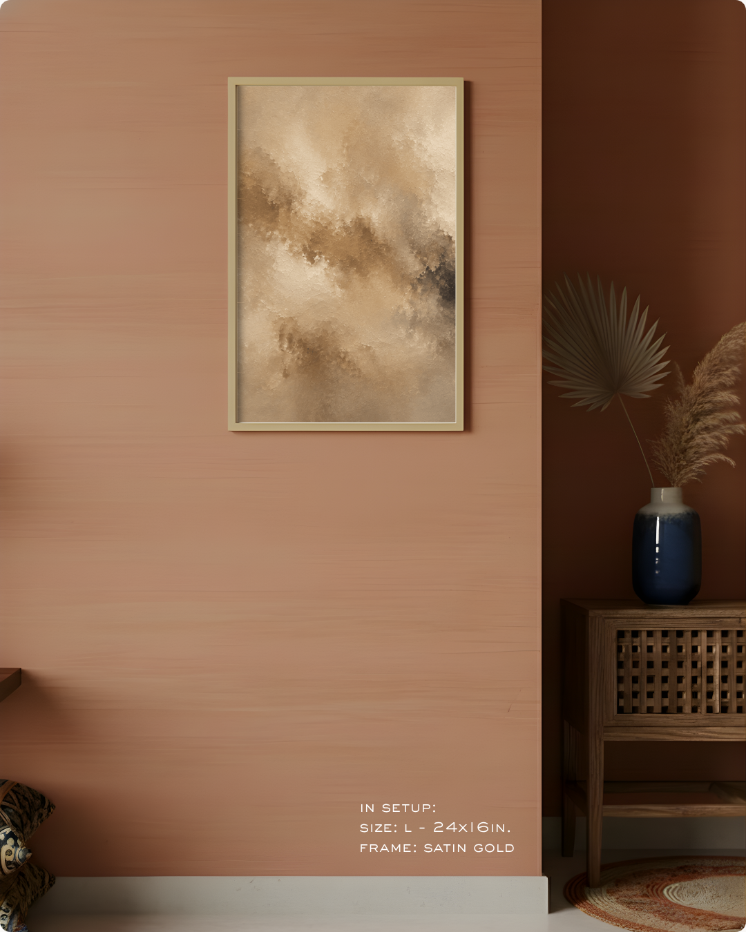 A framed abstract art print with soft beige, brown, and gray tones hanging on a warm terracotta wall in a styled interior setting. The frame is satin gold, and the scene includes a wooden side table, a blue vase with dried palm leaves, and small text at the bottom indicating material and frame details.