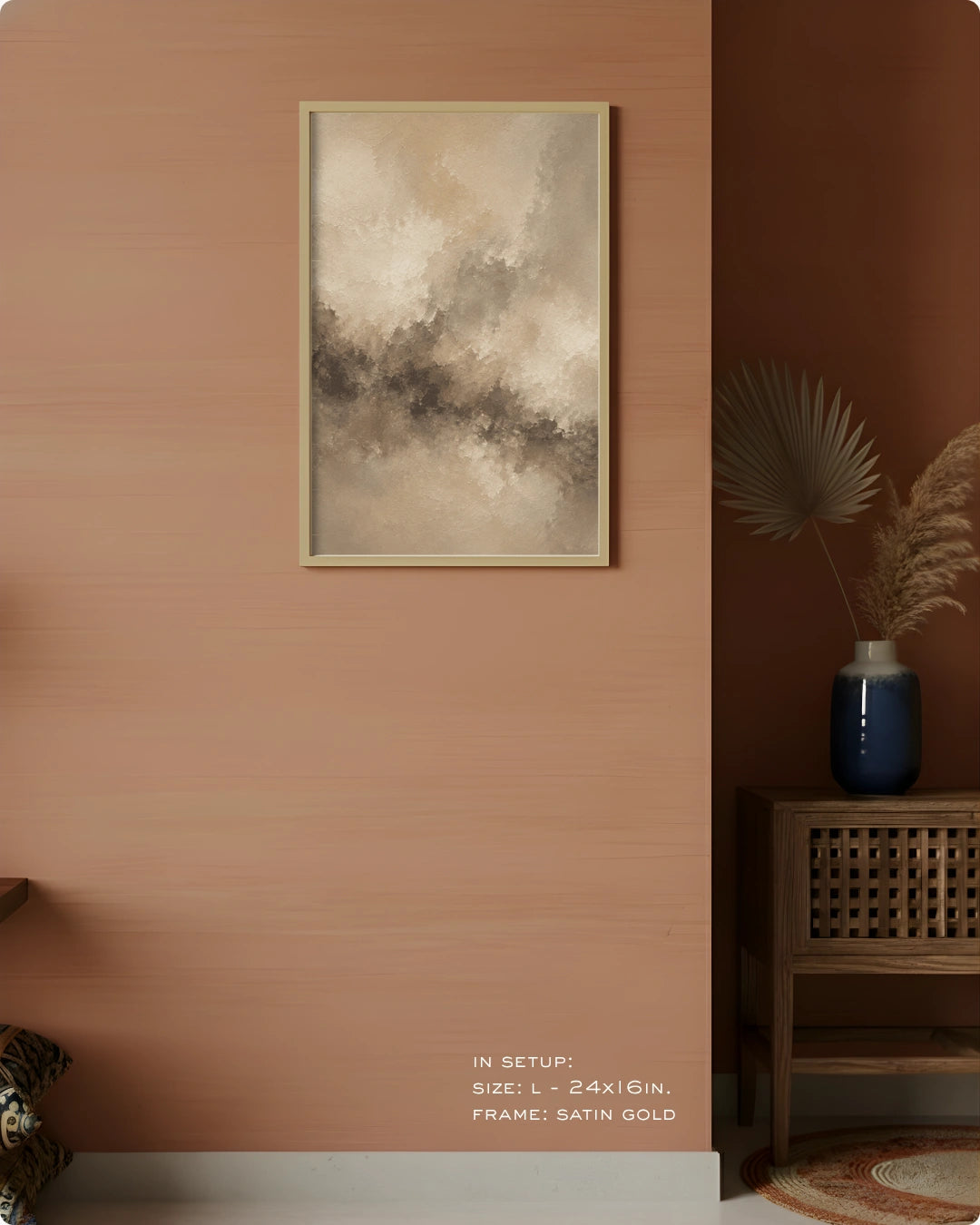 A framed abstract wall art print hangs on a warm peach-pink wall in a styled interior. The artwork features soft beige, cream, and gray brushy tones with a satin gold frame.