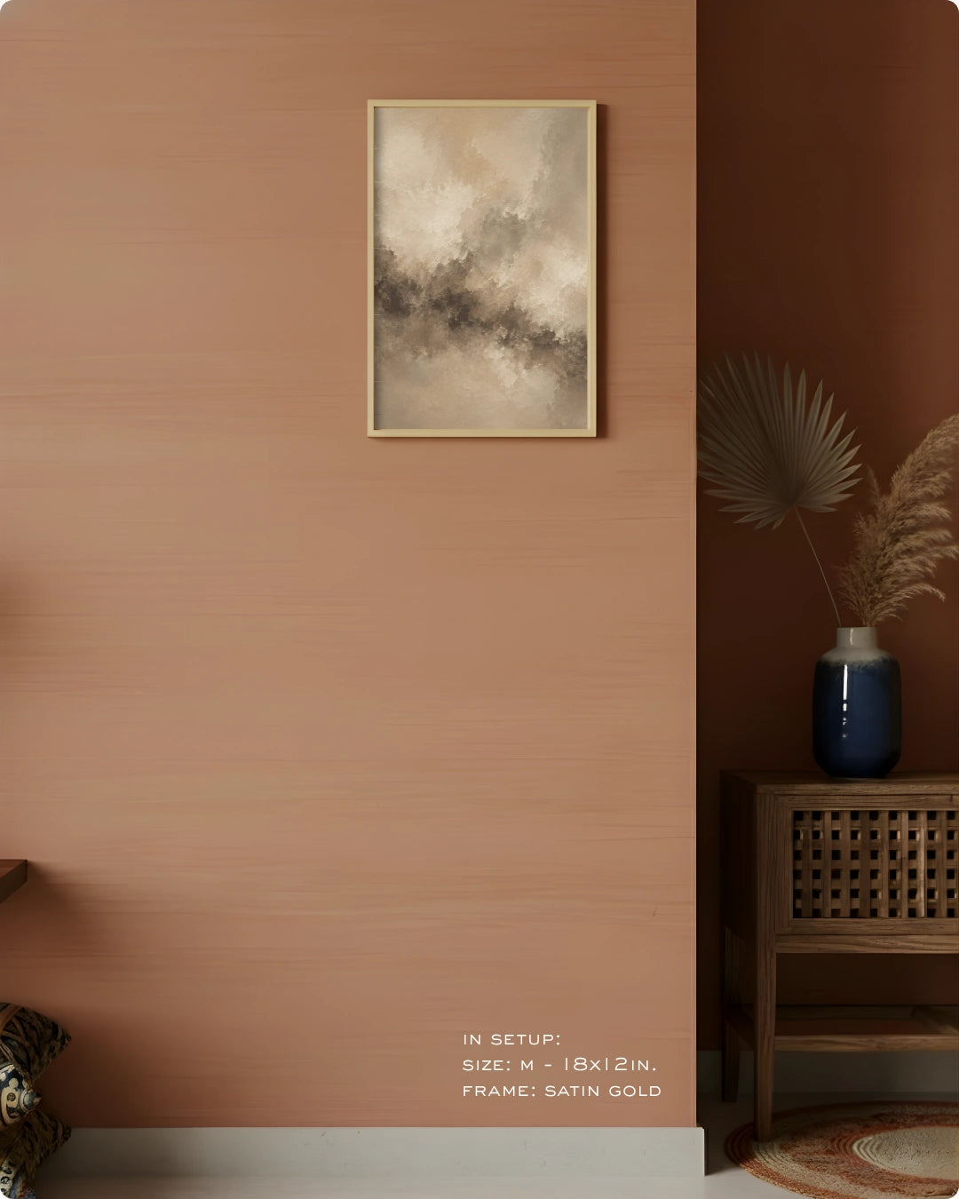 A framed abstract art print with a satin gold frame hangs on a terracotta-colored wall in a styled interior setting. The print features a textured, cloudy pattern in beige, gray, and brown tones.