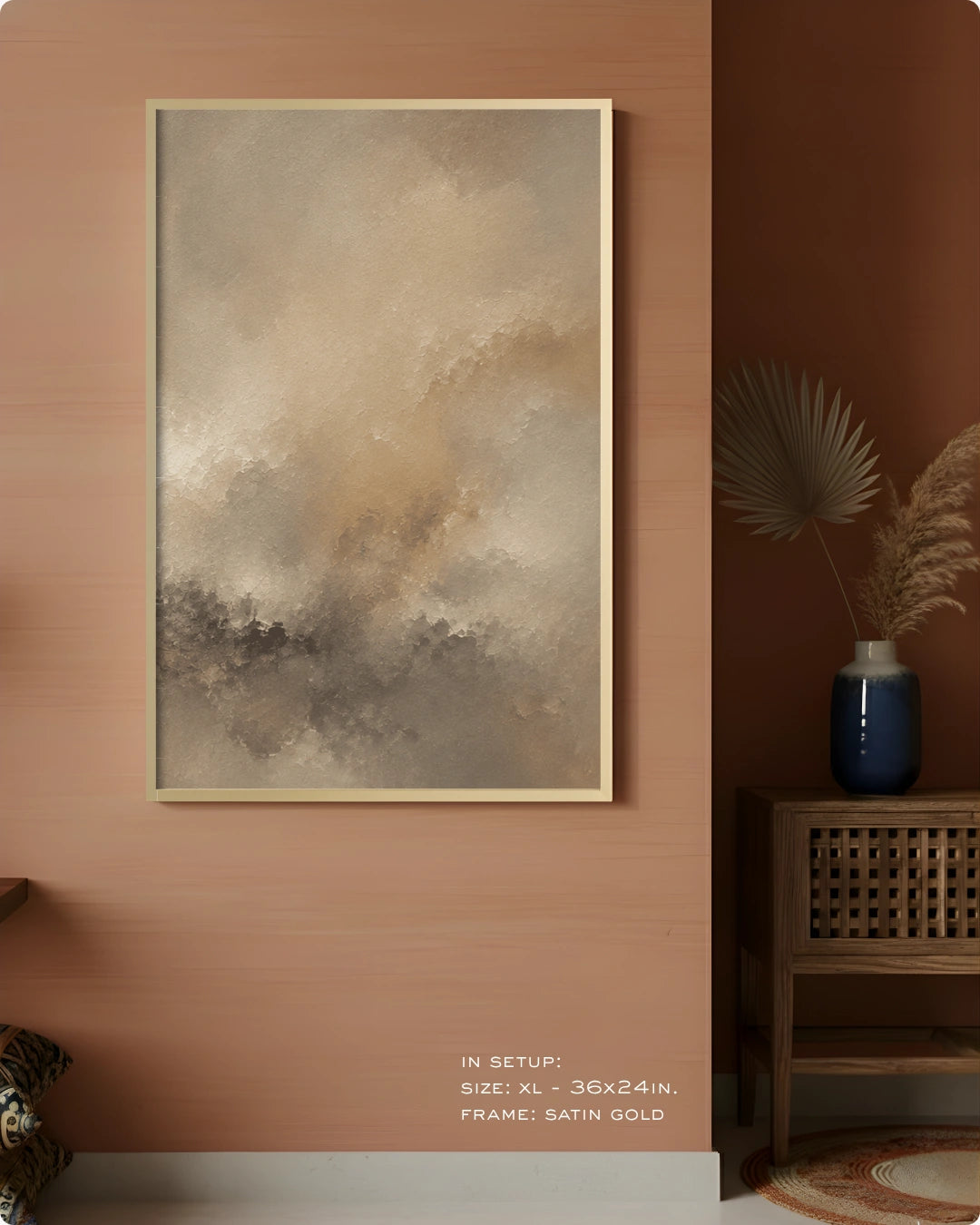 A framed abstract wall art print hangs on a warm terracotta-colored wall in a styled interior. The artwork features soft, painterly clouds and textures in beige, taupe, gray, and brown tones, enclosed in a thin satin gold frame.