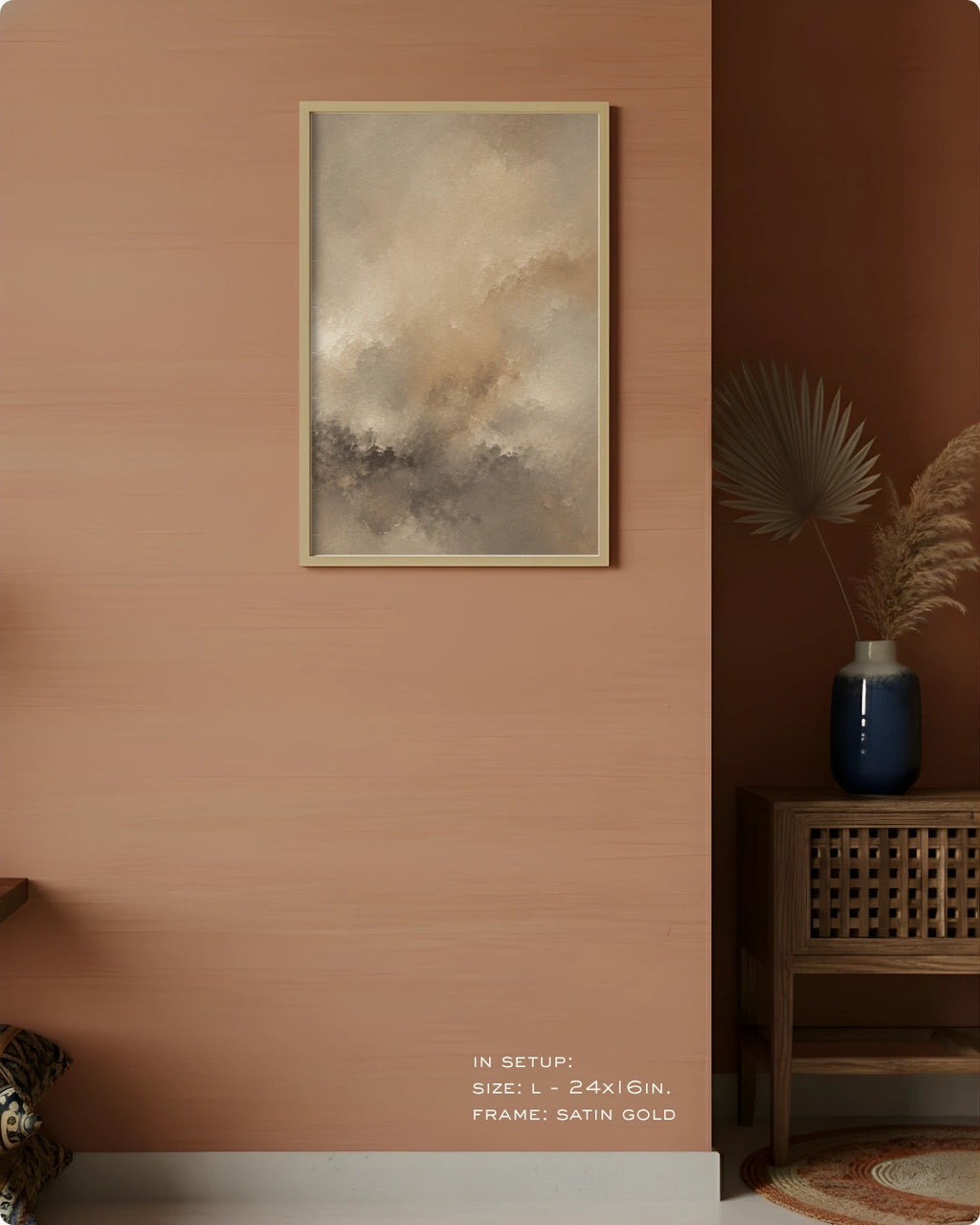 Abstract framed wall art hanging on a warm peach-pink wall in a styled interior, with a wooden side table, blue vase, dried palm leaves, and a small rug visible. The artwork has soft beige, gray, and taupe tones in a satin gold frame.