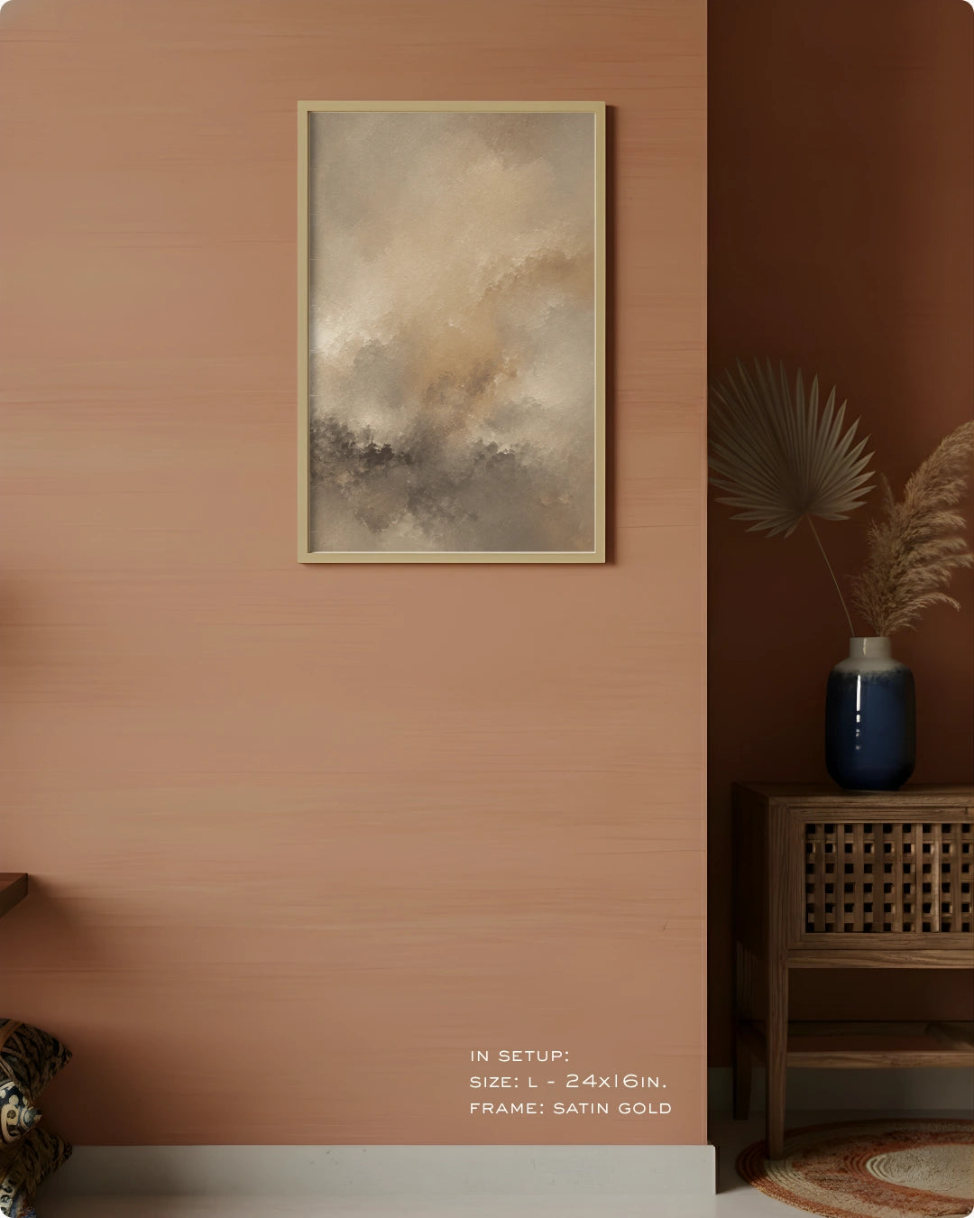 Abstract framed wall art hanging on a warm peach-pink wall in a styled interior, with a wooden side table, blue vase, dried palm leaves, and a small rug visible. The artwork has soft beige, gray, and taupe tones in a satin gold frame.