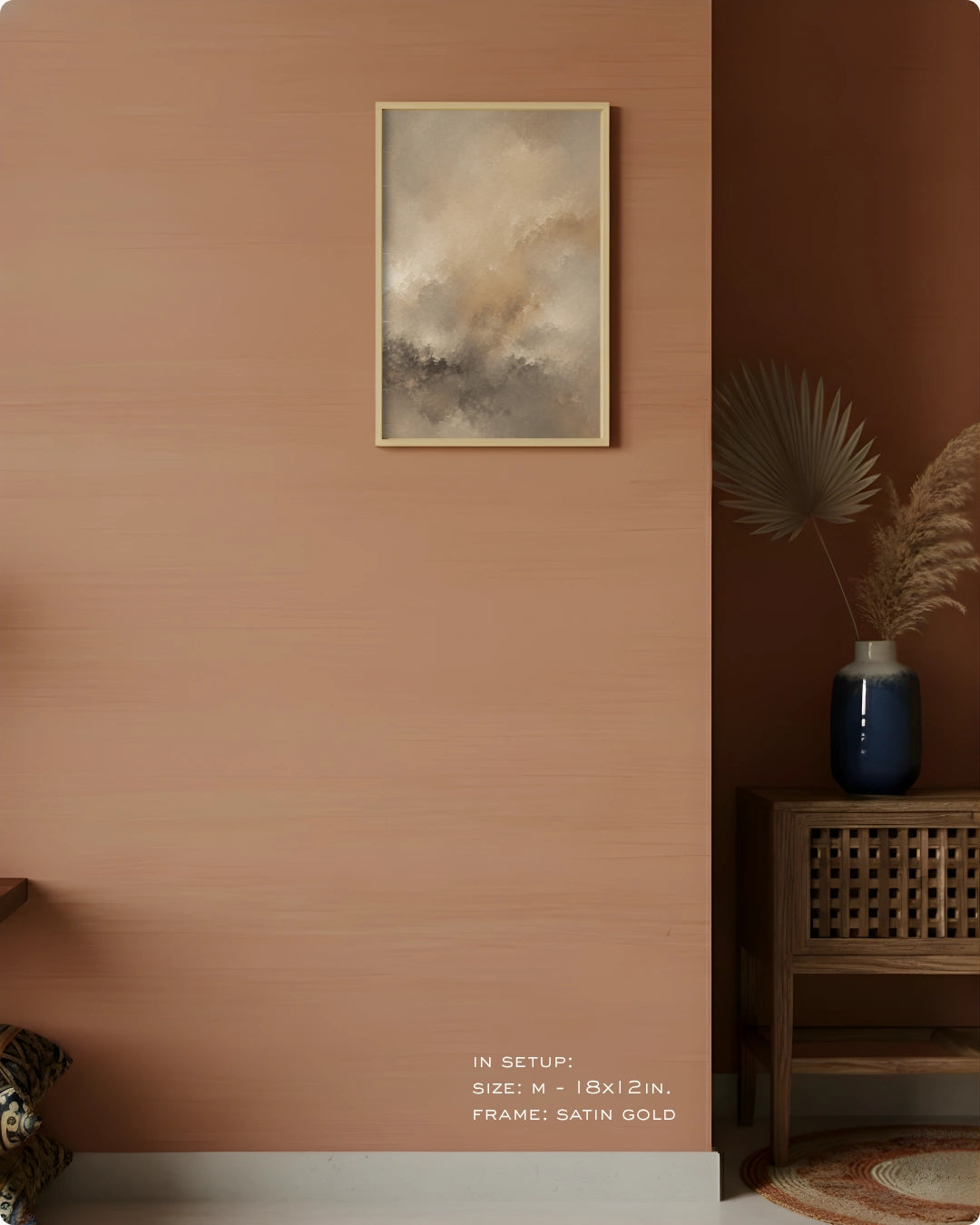 A framed abstract art print with a satin gold frame hangs on a textured terracotta wall in a styled interior setting. The print features soft, painterly clouds and smoke in beige, gray, and brown tones.