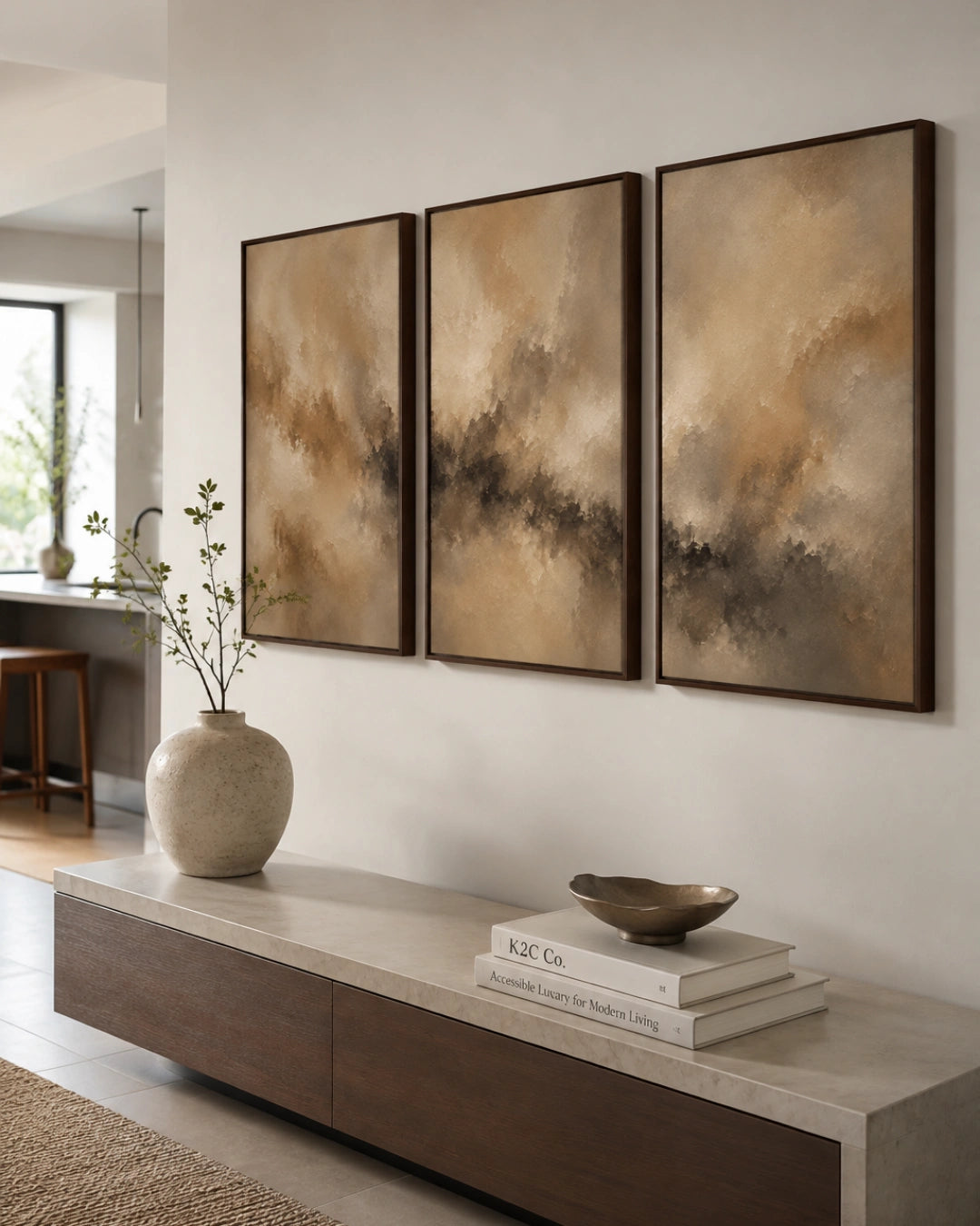 Three framed abstract wall art panels in warm beige, brown, and charcoal tones hang above a long wood console in a modern interior with a vase and books.