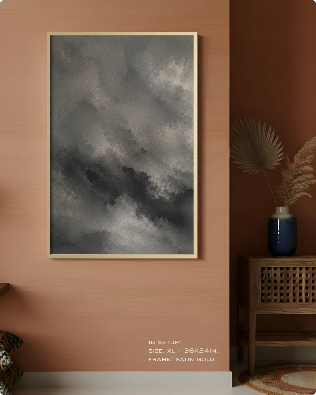 A framed wall art print featuring a dramatic gray and black cloudy sky, displayed on a warm terracotta wall in a styled interior setting with a wooden side table, blue vase, and dried palm leaves.
