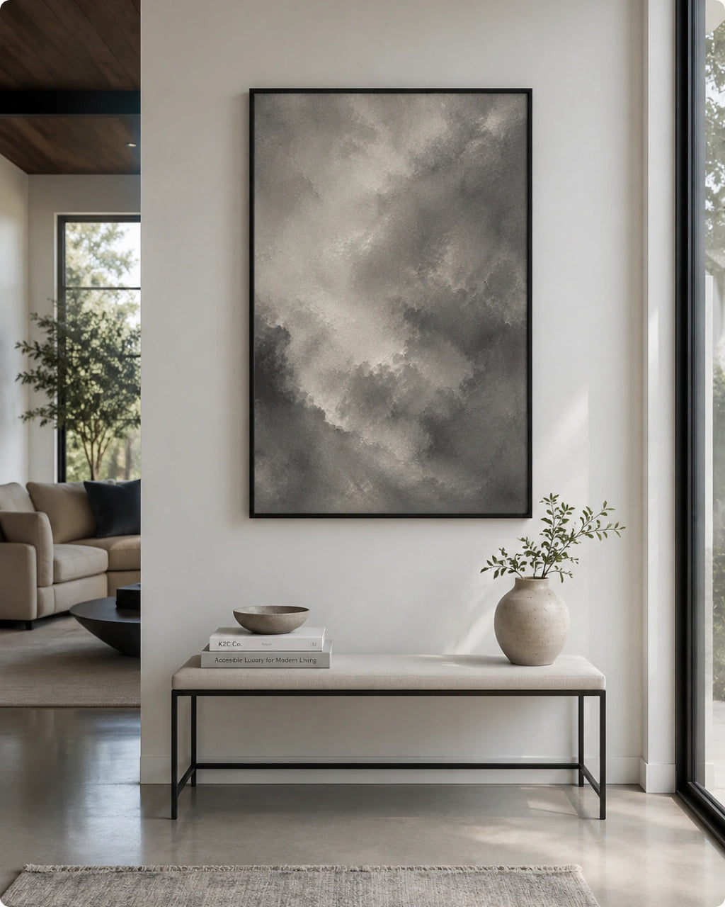 A large vertical framed wall art print featuring a gray and white cloudy abstract scene, displayed in a modern living room setting above a light-colored bench with a vase and books.