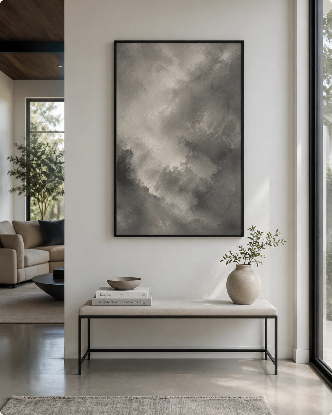 A large vertical framed wall art print featuring a gray and white cloudy abstract scene, displayed in a modern living room setting above a light-colored bench with a vase and books.