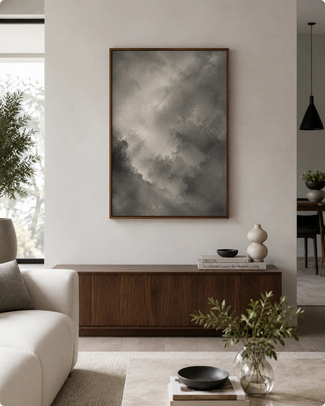 A framed vertical art print featuring a dramatic gray and white cloudy sky, displayed on a light beige wall above a dark wood console table in a modern living room setting.