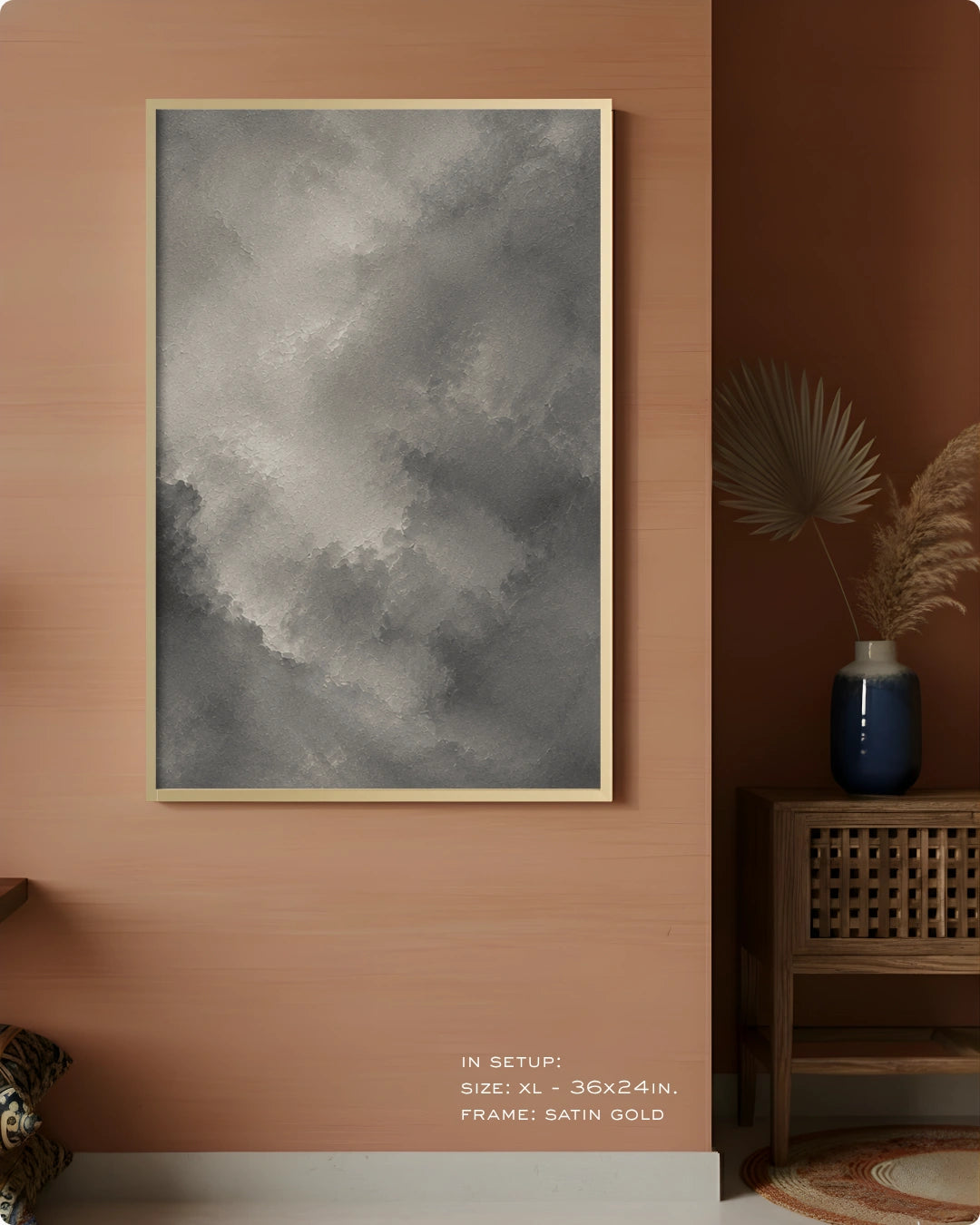 A framed wall art print featuring a gray and white textured cloudy or smoky abstract design, hung on a warm peach-colored wall in a styled interior setting with a side table and vase.